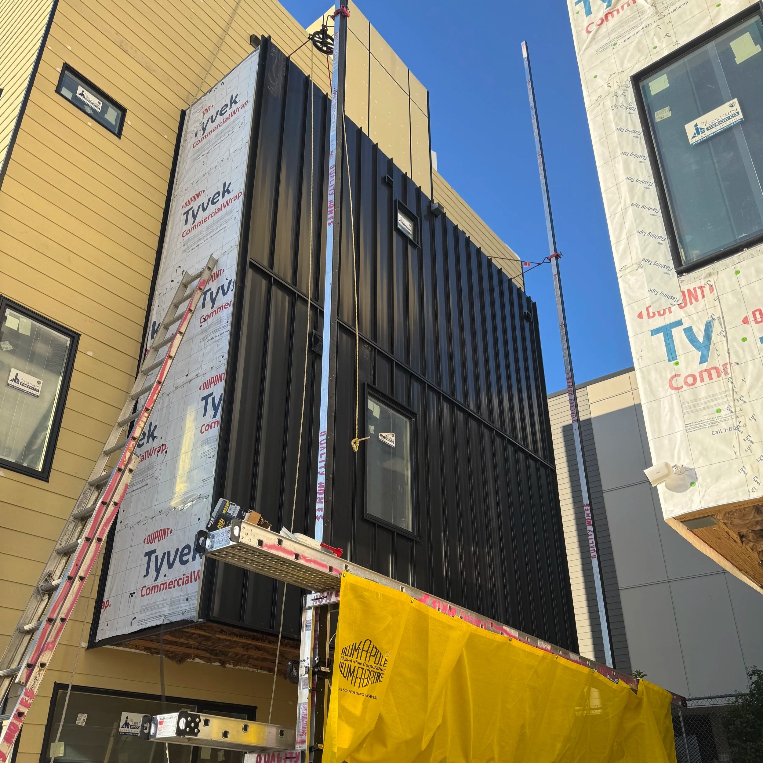 Construction site with building exterior under renovation, showing partially installed black siding, yellow wrapping, a ladder, and a crane against a clear blue sky.