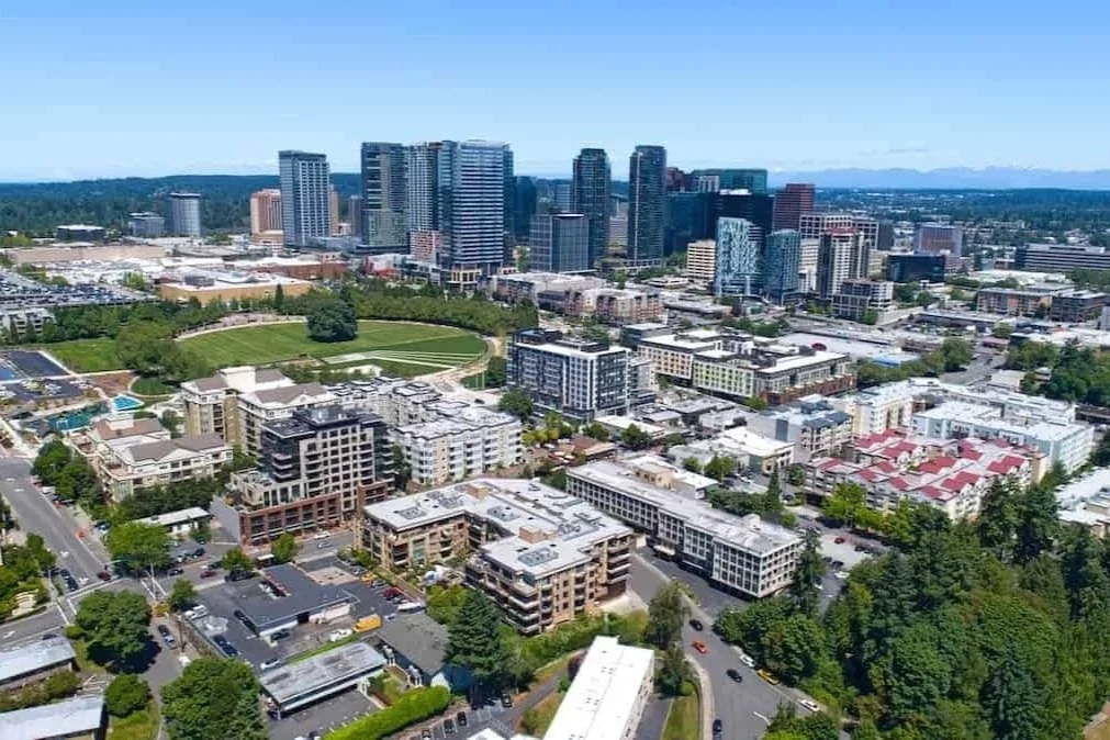 Aerial view of a cityscape with high-rise buildings, residential complexes, and a park with a soccer field, under a clear blue sky.