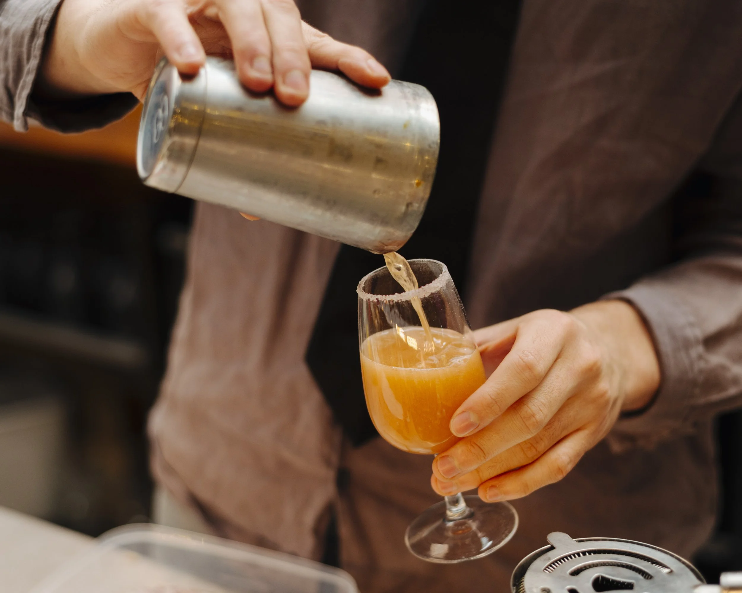 Person pouring a cocktail or beverage from a shaker into a glass with salted rim, in a bar or restaurant setting.