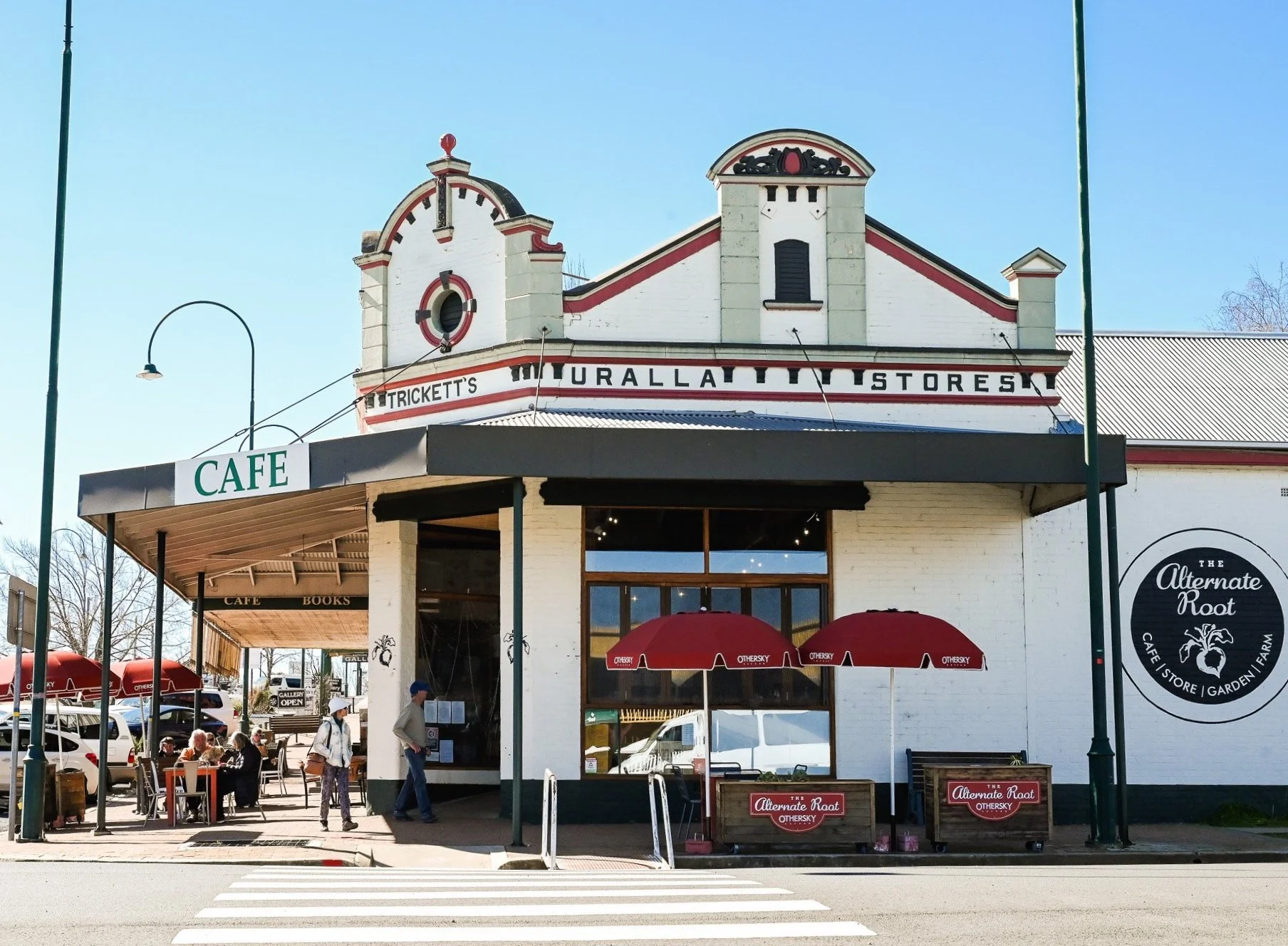 Exterior of a quaint café and bookstore named The Alternate Root, with outdoor seating and red umbrellas, on a sunny day.