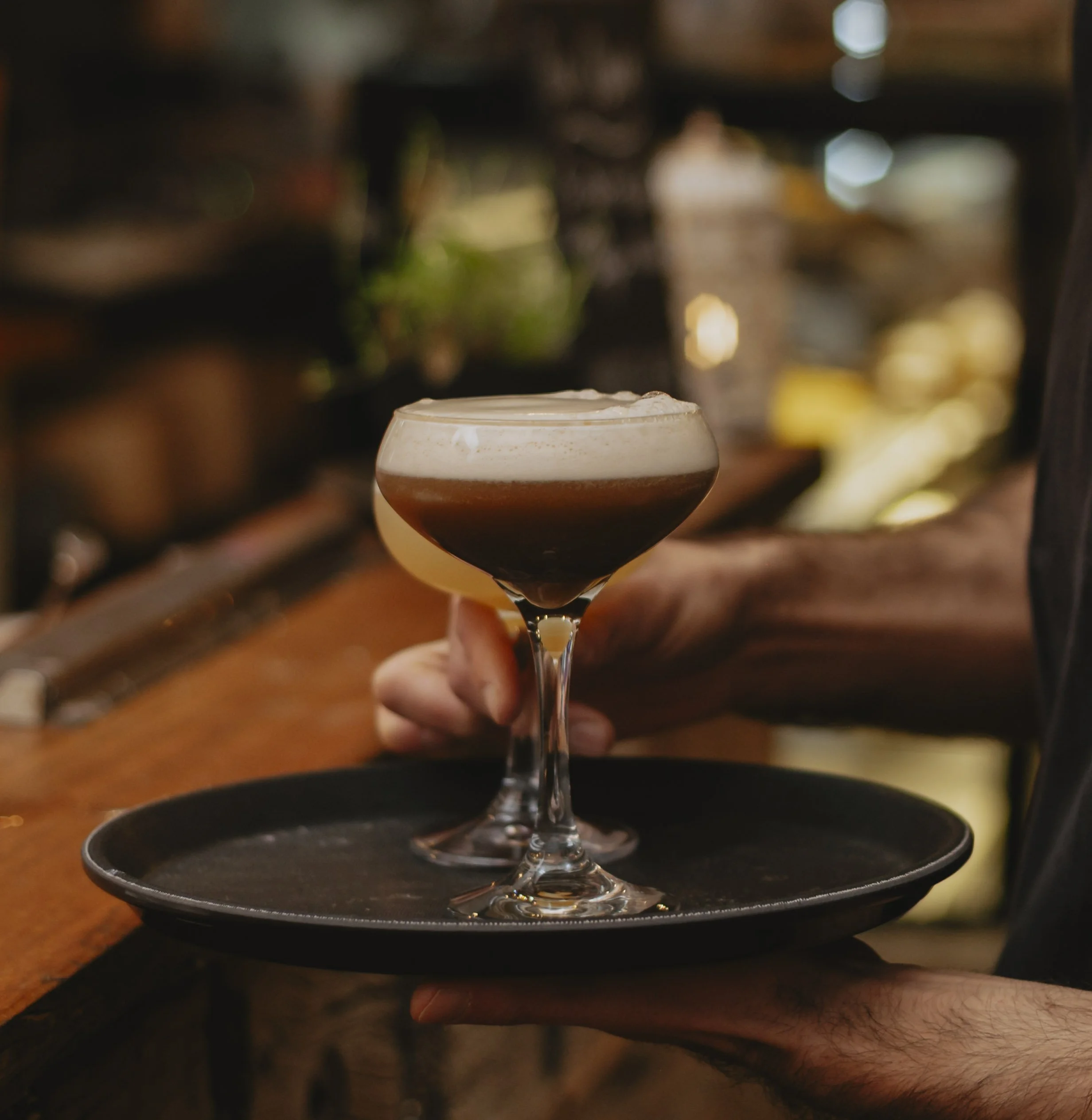 A bartender holding a black tray with two cocktail glasses, one with a layered drink and the other with a dark brown drink topped with foam, at a bar.