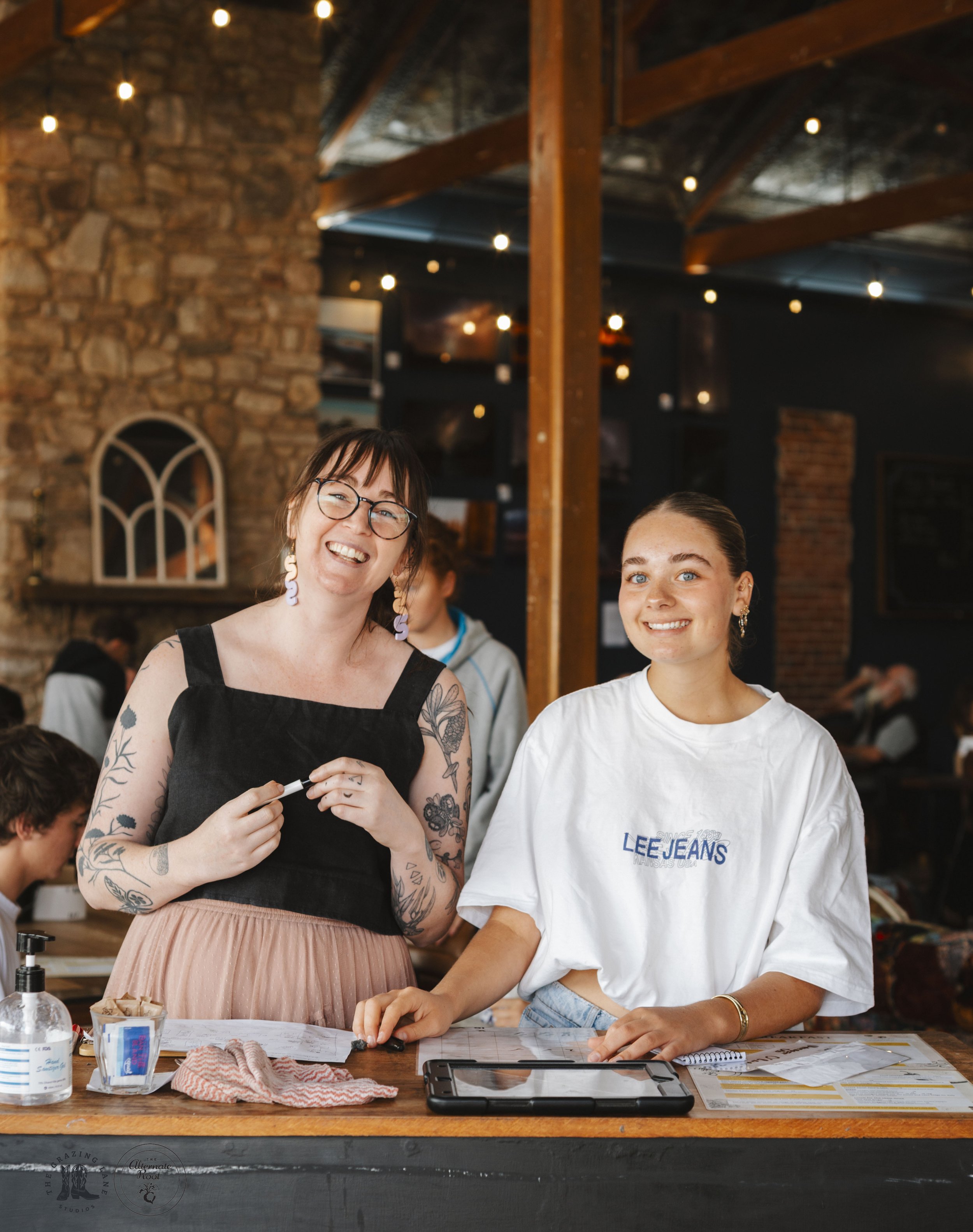 Two smiling women behind a restaurant or cafe counter with various papers, a tablet, hand sanitizer, and a cloth on the counter, in a warm, rustic interior with wooden beams and brick walls.