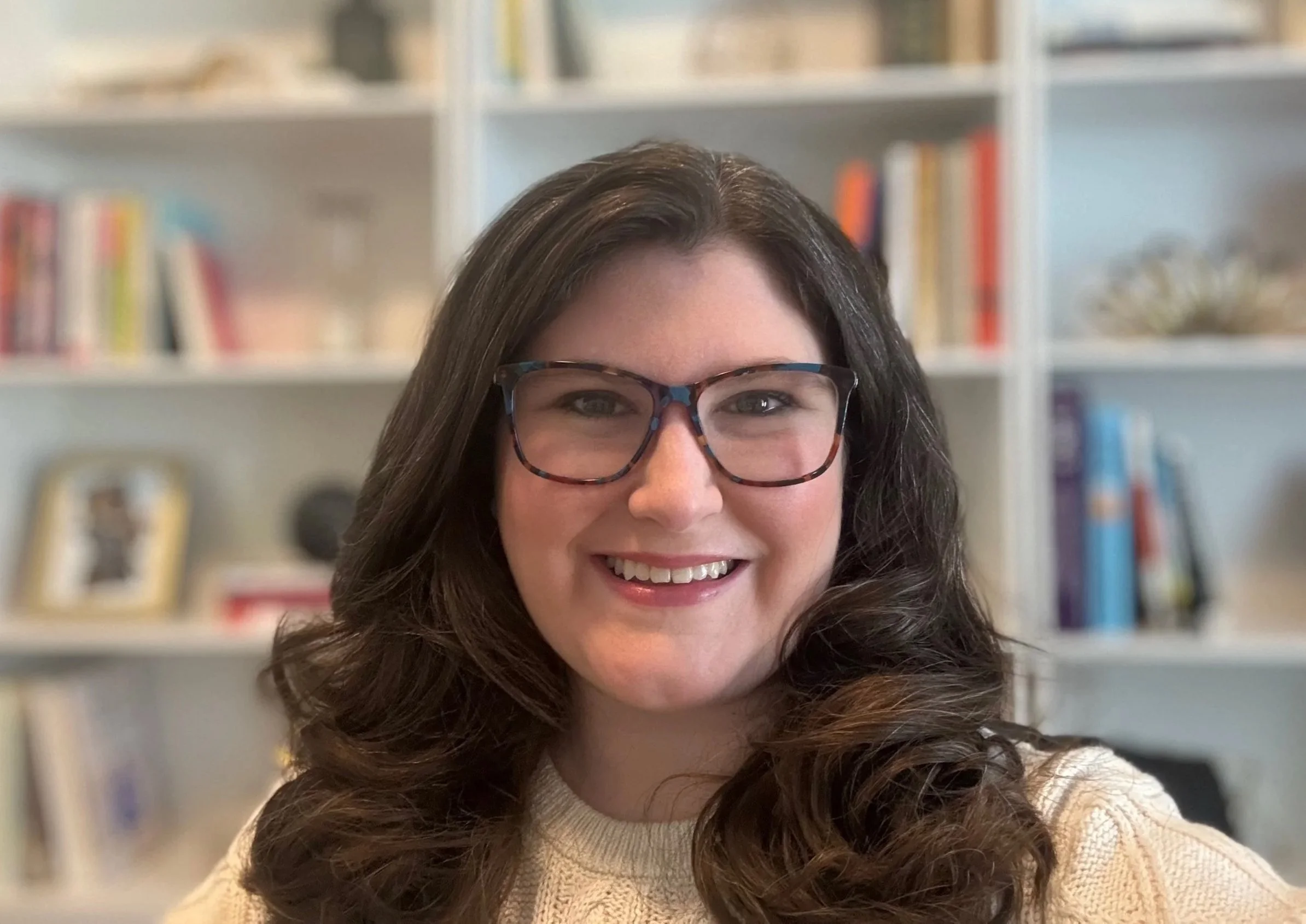 A headshot of Dr. Bethany Rothamel, shown smiling in front of a blurred bookshelf filled with books and decorative items.