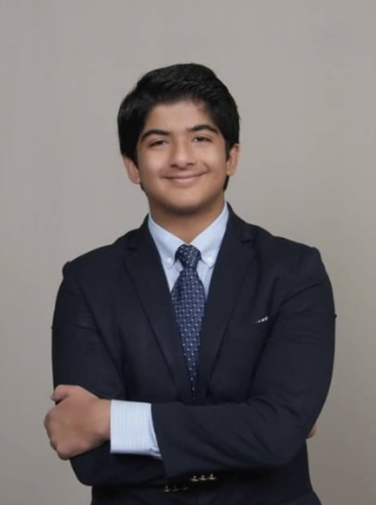 Young man with short black hair, wearing a dark suit, light blue dress shirt, and patterned tie, standing with arms crossed and smiling against a plain gray background.