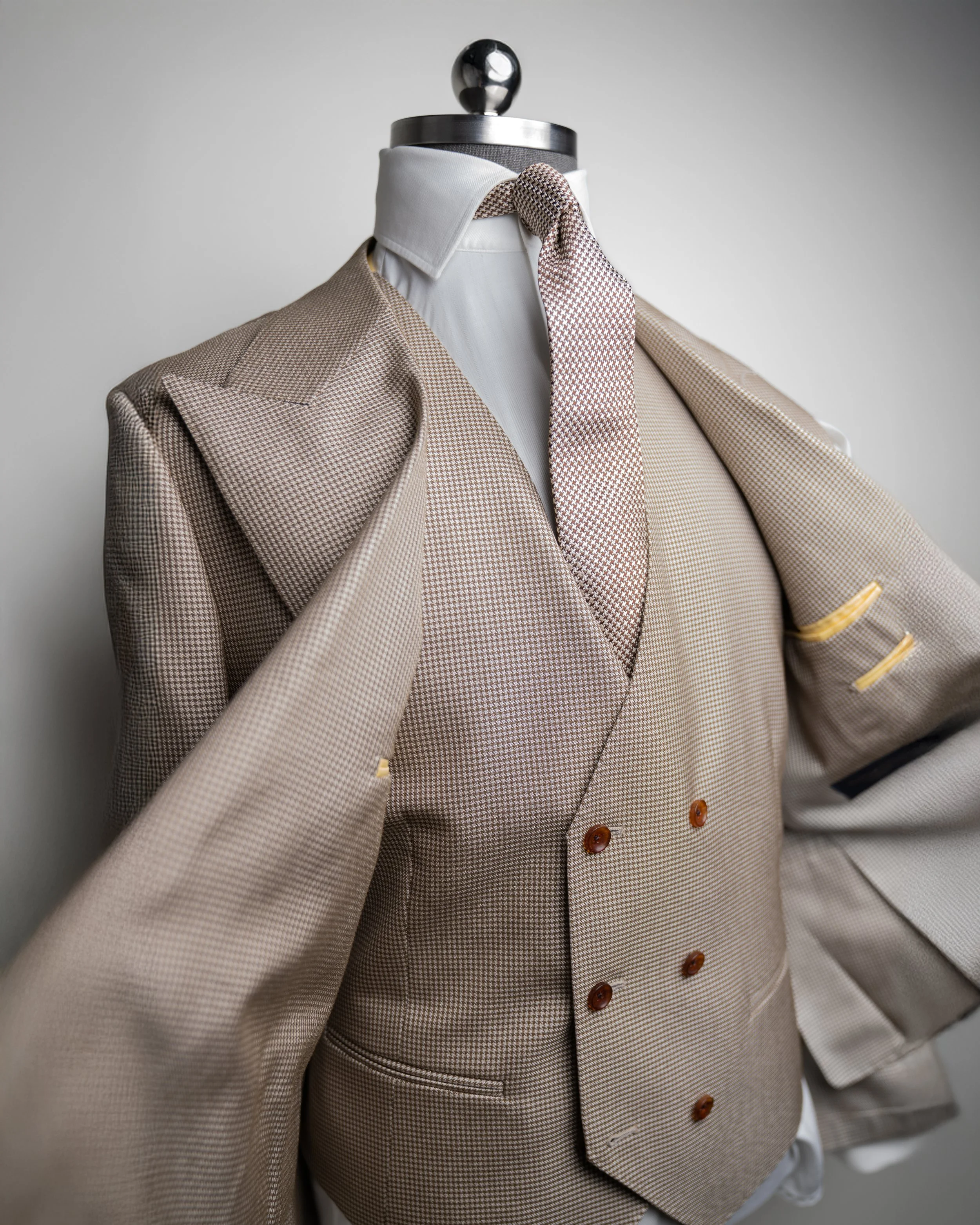A mannequin dressed in a beige houndstooth blazer over a white shirt with a bundled matching tie, against a plain gray background.