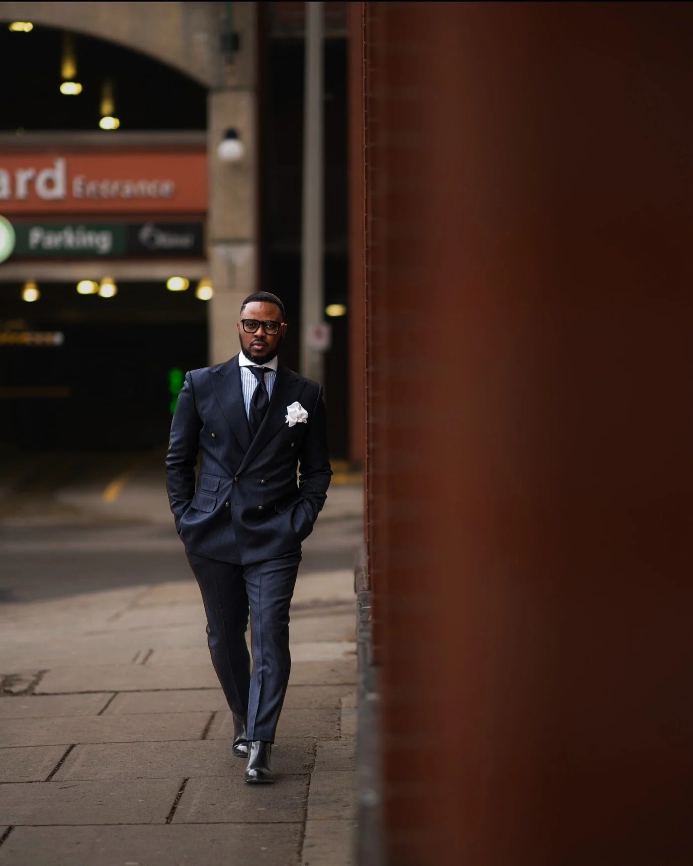A man in a navy blue pinstripe suit with a white pocket square, tie, and glasses walking on a city sidewalk during daytime, with parking garage signs in the background.