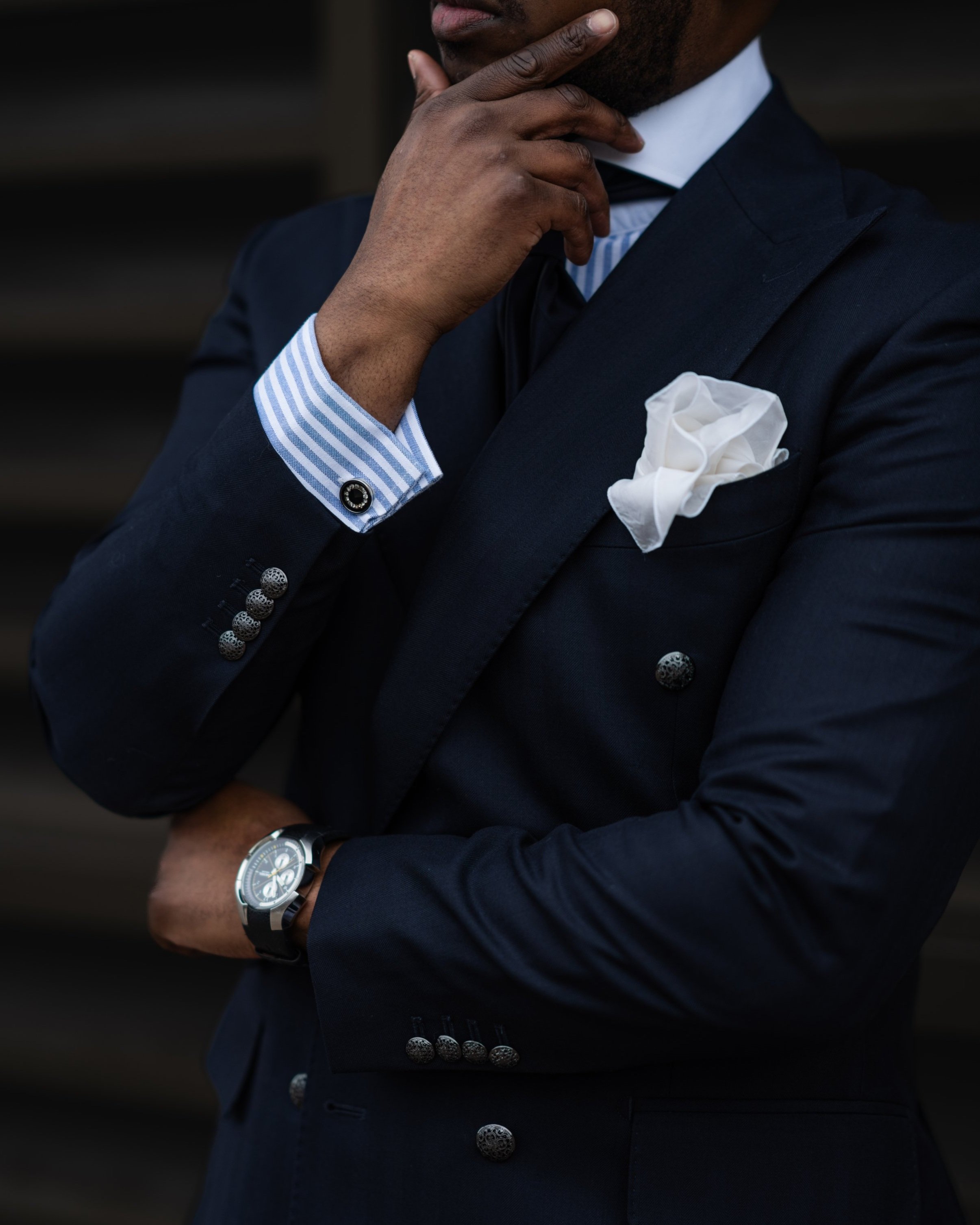 Close-up of a man in a navy blue tuxedo with a white pocket square, wearing a wristwatch, touching his chin, with a dark background.
