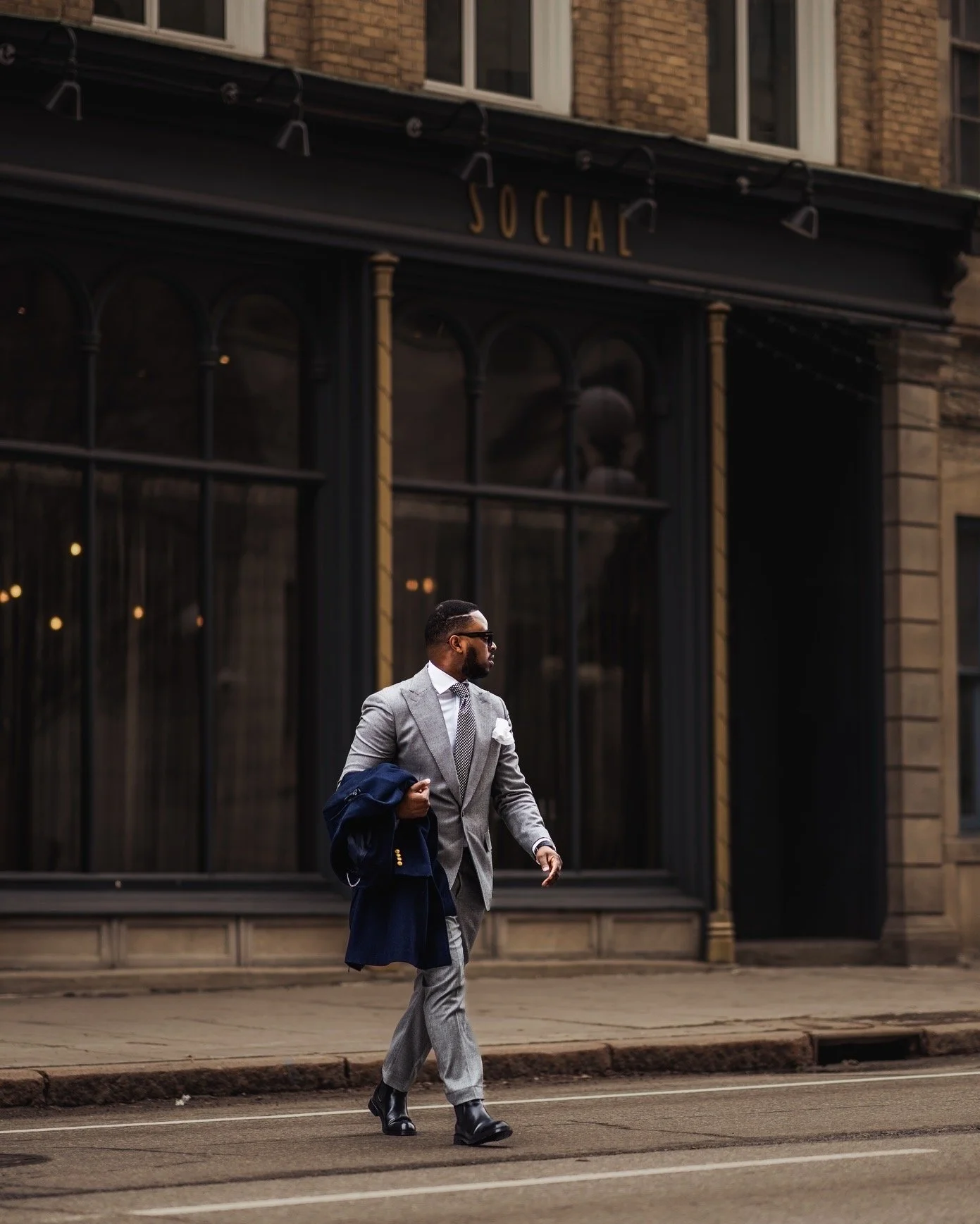 A man in a gray suit with a white shirt walking on the sidewalk in front of a building with black and gold details and large windows. He is carrying a dark jacket and wearing sunglasses.