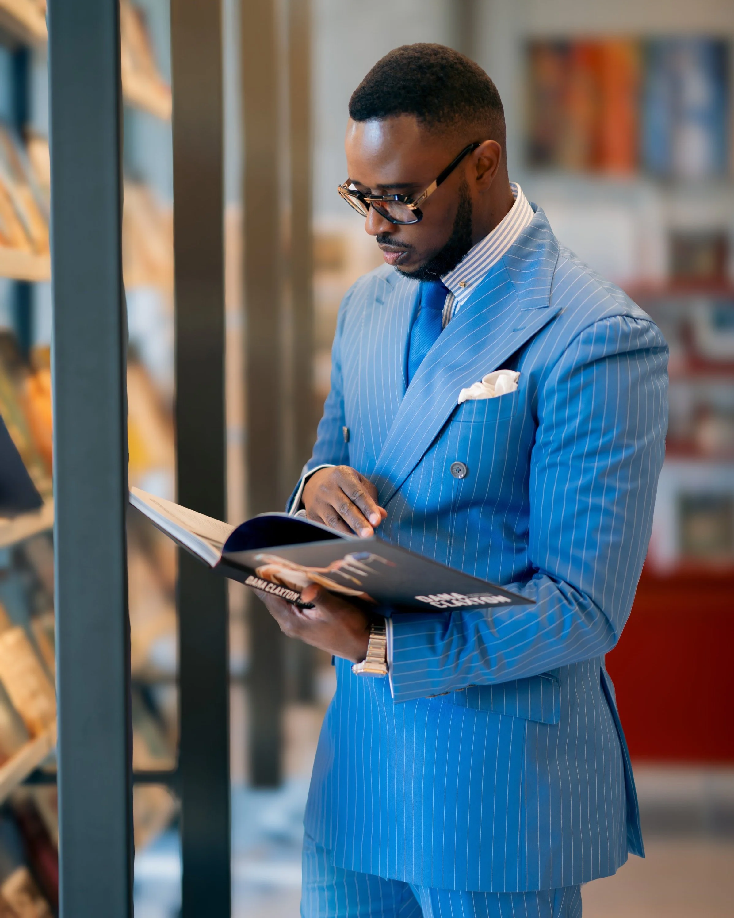 A man in a blue pinstripe suit, wearing glasses, reading a magazine or brochure in a bookstore or library.