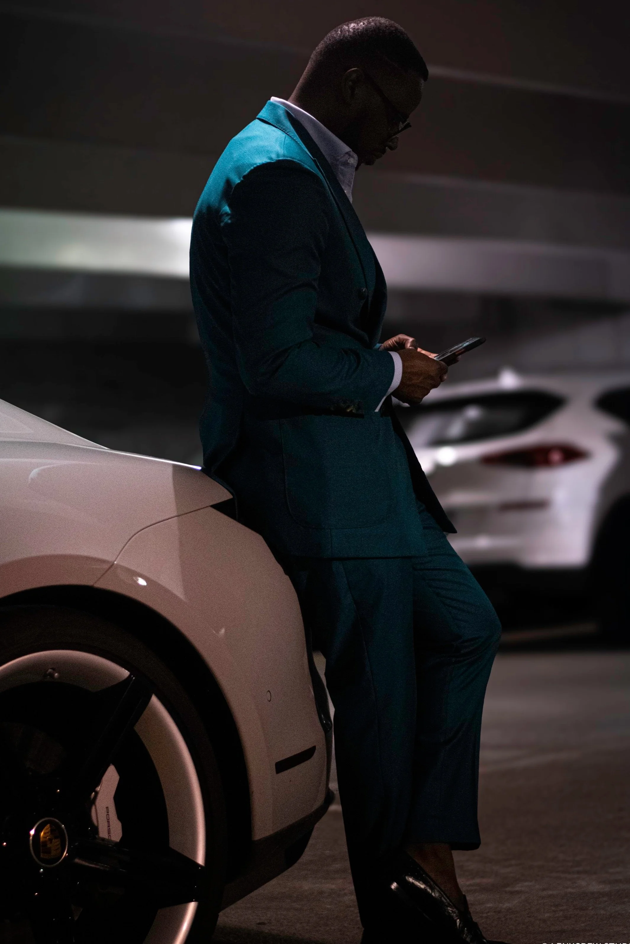 A man in a suit leaning against a car in a dimly lit parking garage, looking at his phone.