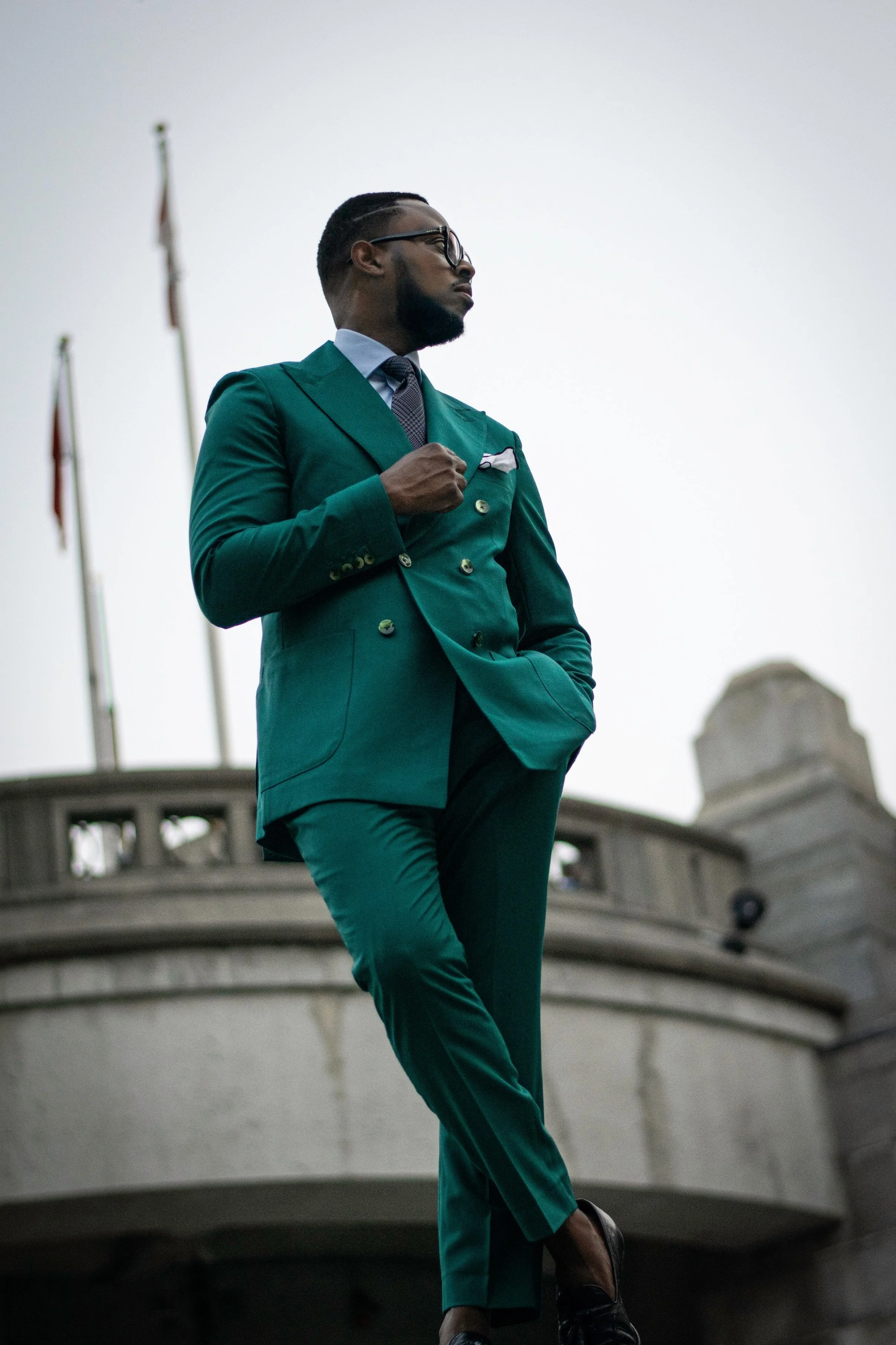 A man in a teal double-breasted suit, white shirt, and tie standing outdoors near a historic building with flags, looking away and holding his jacket lapel, with a cloudy sky in the background.
