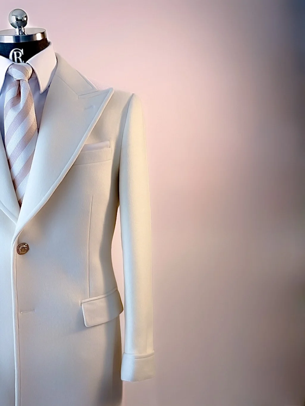 A close-up of a mannequin dressed in a cream-colored tuxedo with a pink and white striped tie and a white shirt, against a plain background.
