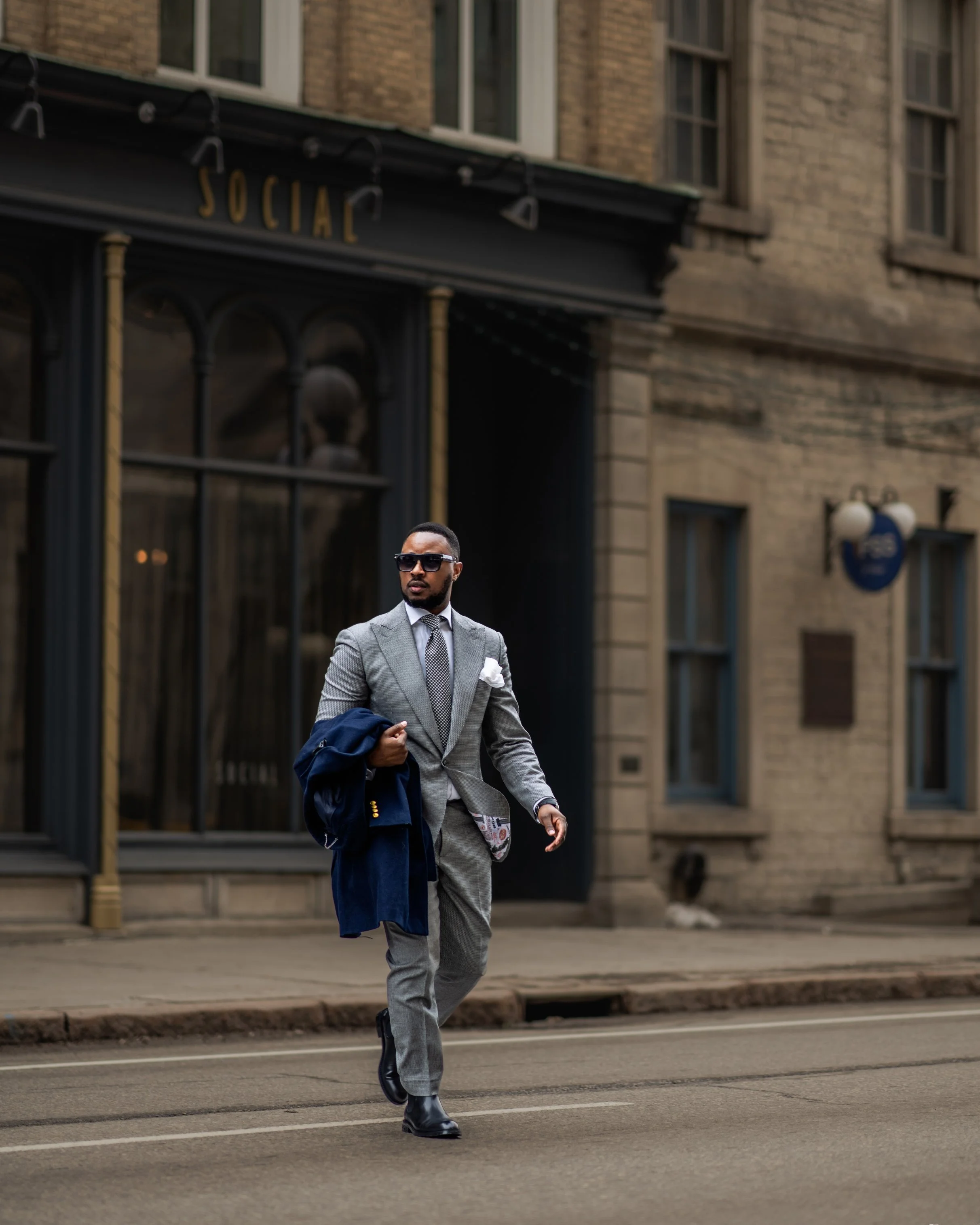 A stylish man in a gray suit with a patterned tie and white pocket square walking on a city street, carrying a navy blue jacket, wearing sunglasses, and black boots.
