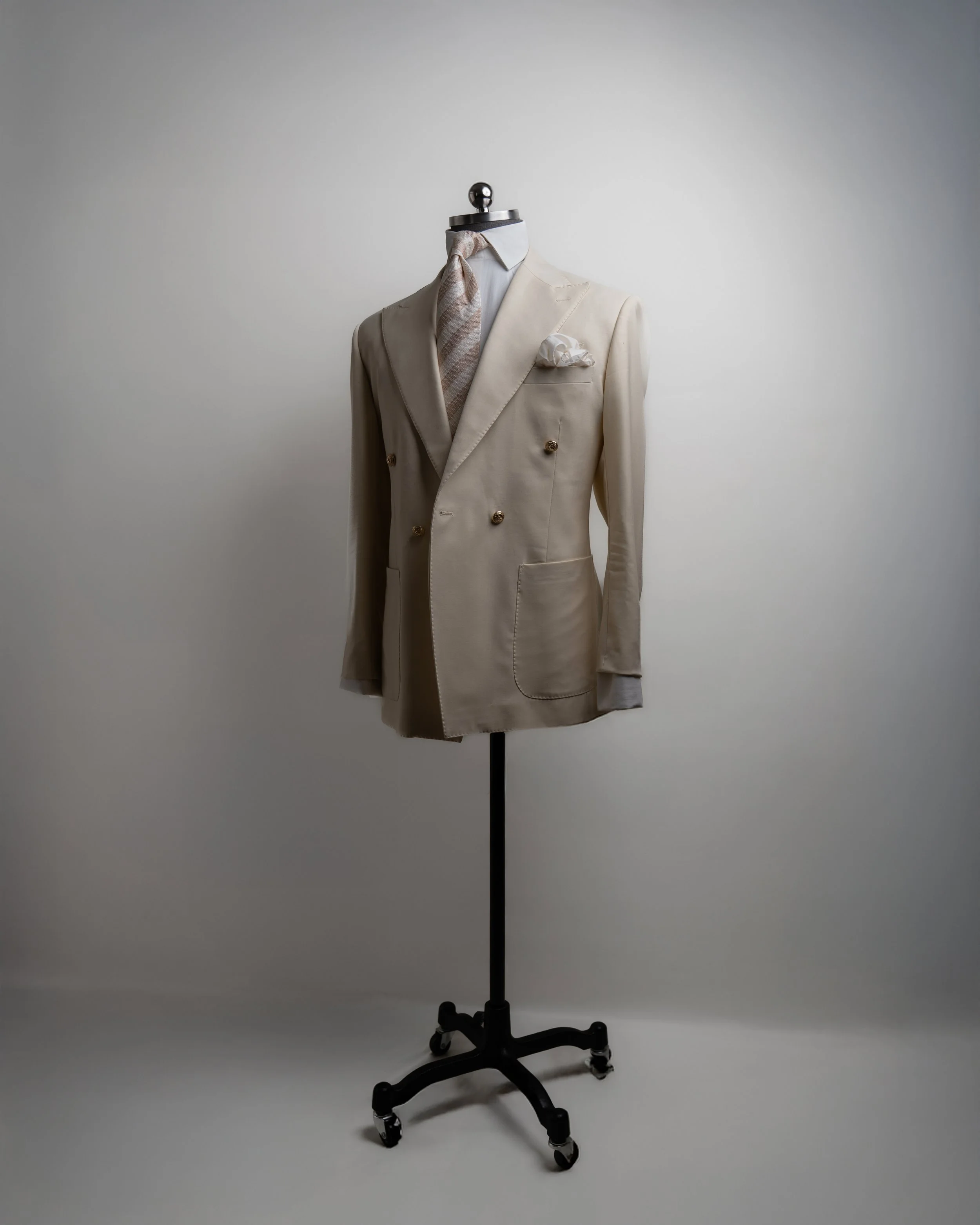 A cream-colored tuxedo with gold buttons on a mannequin, decorated with a white pocket square, a brown and beige striped tie, and a white shirt, on a black stand against a plain background.