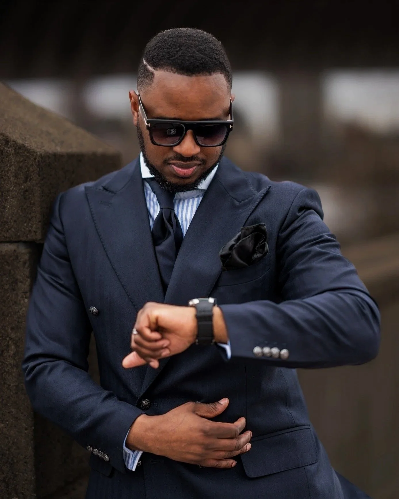 A man in a navy suit with a striped shirt and tie, wearing sunglasses, looking at his wristwatch outdoors.