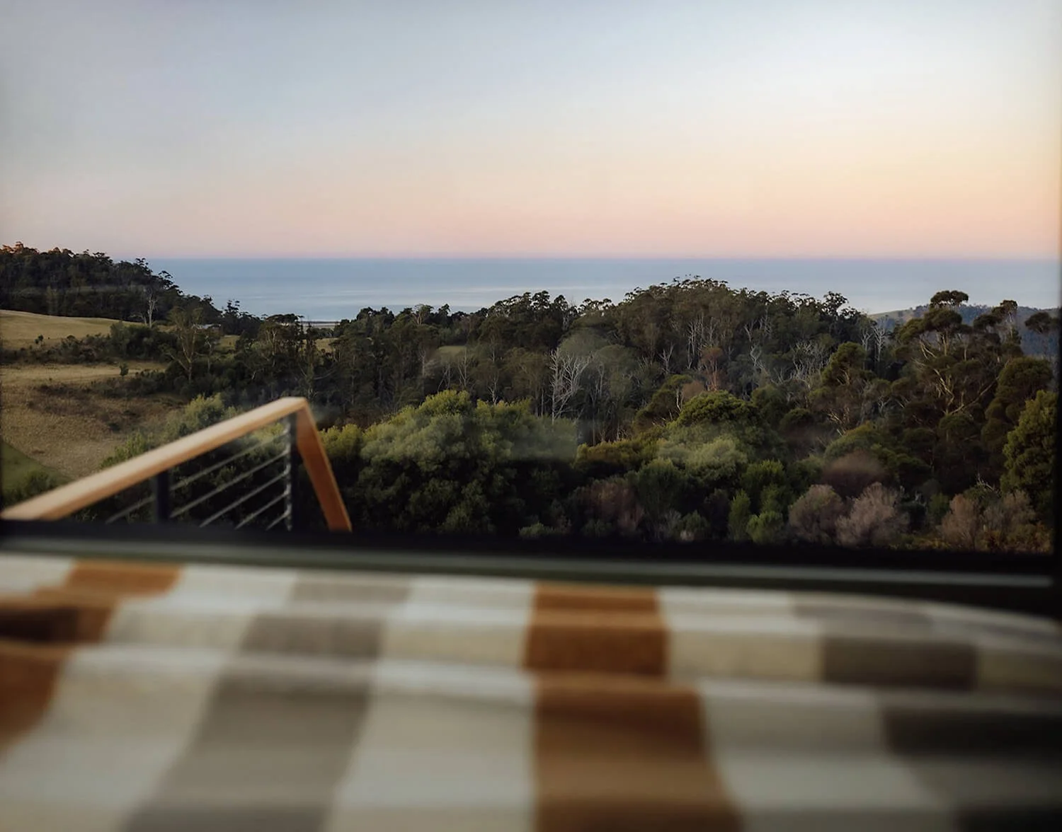 View of a scenic landscape with trees, hills, and the ocean in the distance, viewed from inside a house or cafe, with a checkered table in the foreground.