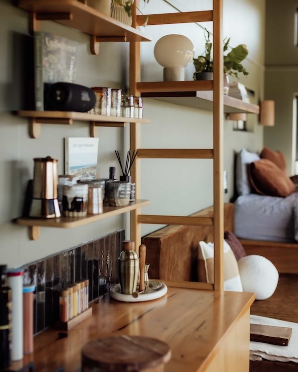 Photo of a cozy living room corner featuring a wall with green paint and wooden shelves holding books, plants, and decorative items. A wooden sideboard with a metallic pitcher and other accessories is in front. In the background, a bed with pillows is visible.