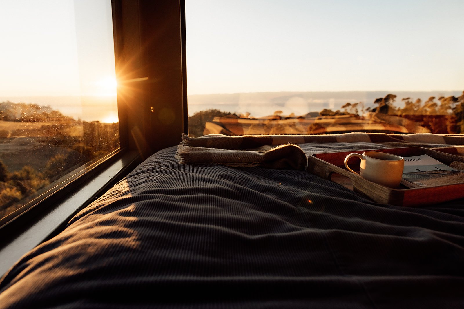 Sunrise view from a bed with a tray holding a coffee mug and a magazine near a window showing trees and a body of water.