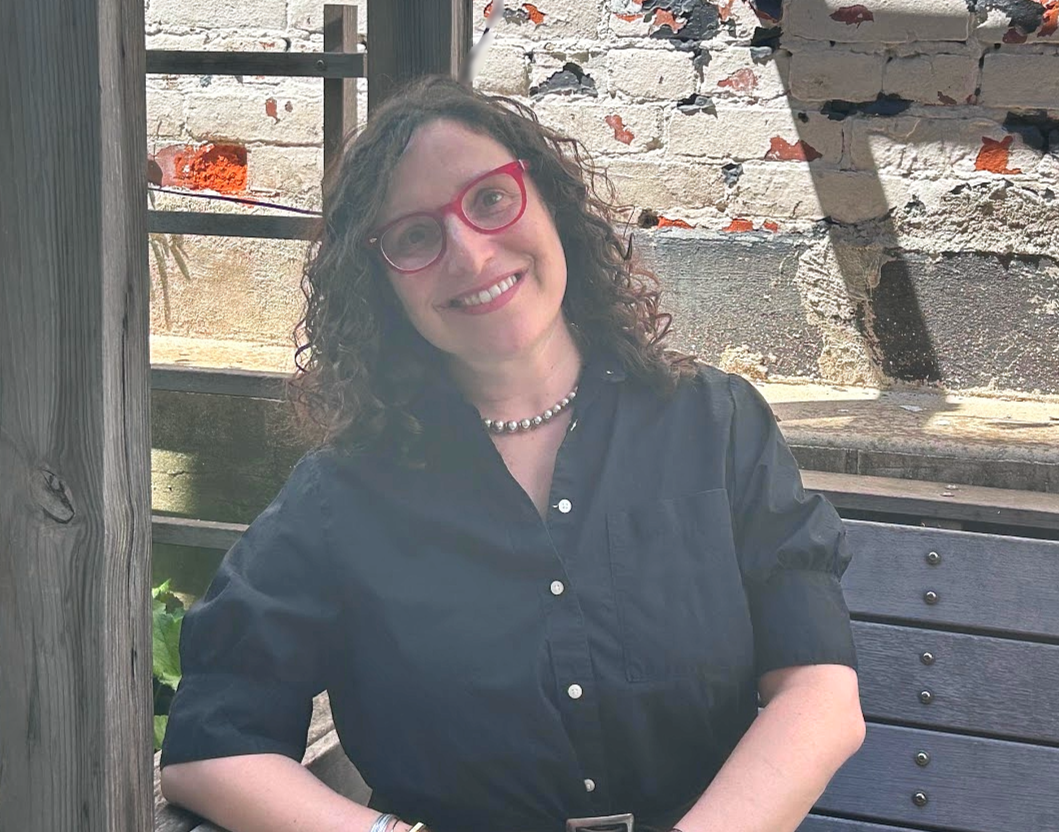 This is a photo of a smiling, White-presenting person with shoulder-length, curly brown hair and red glasses wearing a black button up and necklace outside in front of a brick background.