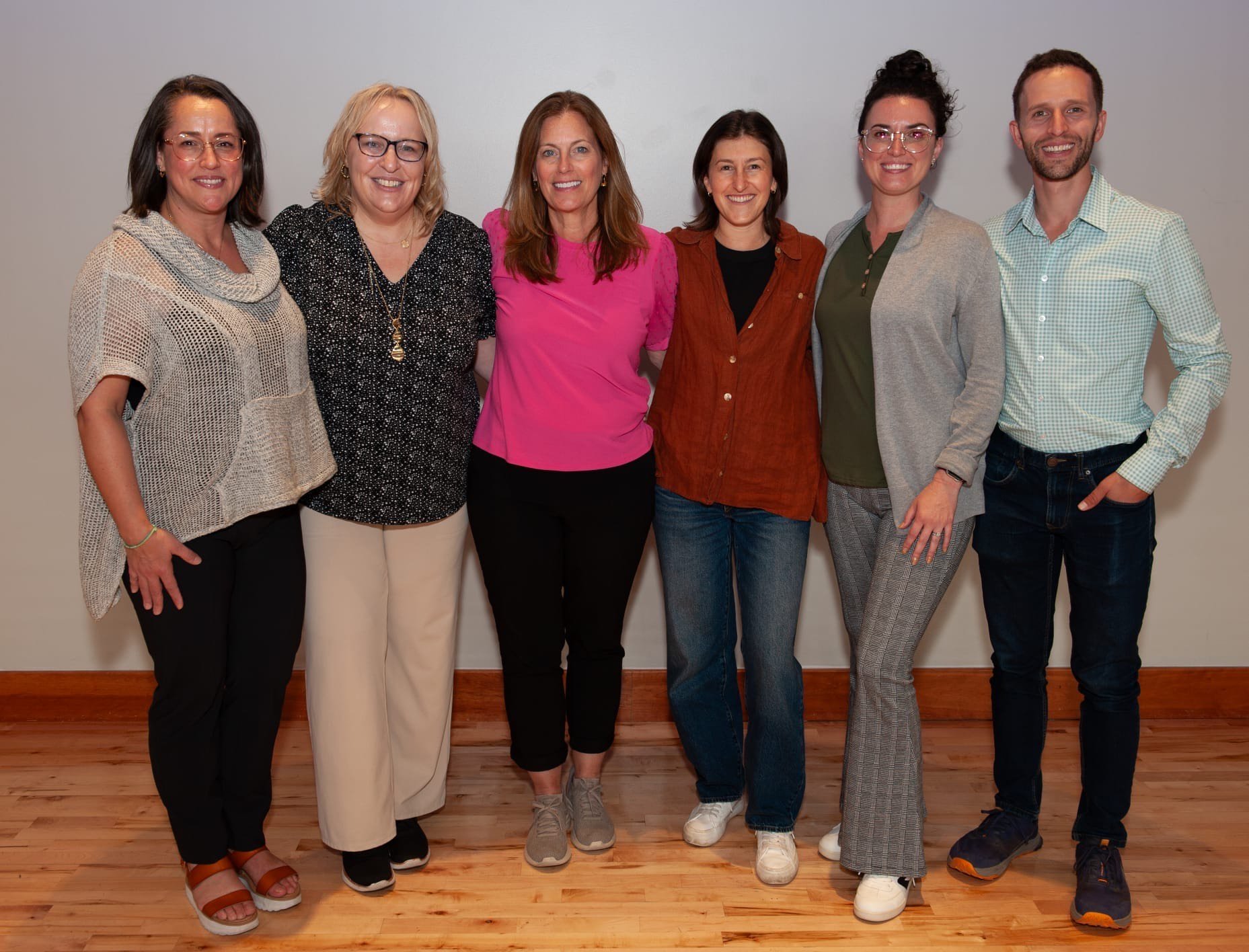 Five women and one man standing side by side in front of a plain wall, all smiling at the camera. They are dressed in casual to business casual attire, on a wooden floor.