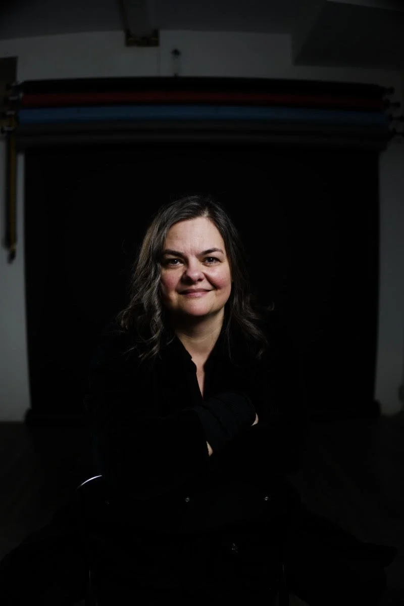 Portrait of a middle-aged man with a bald head, wearing a light blue button-up shirt, smiling in front of large windows with an outdoor view of trees and buildings.