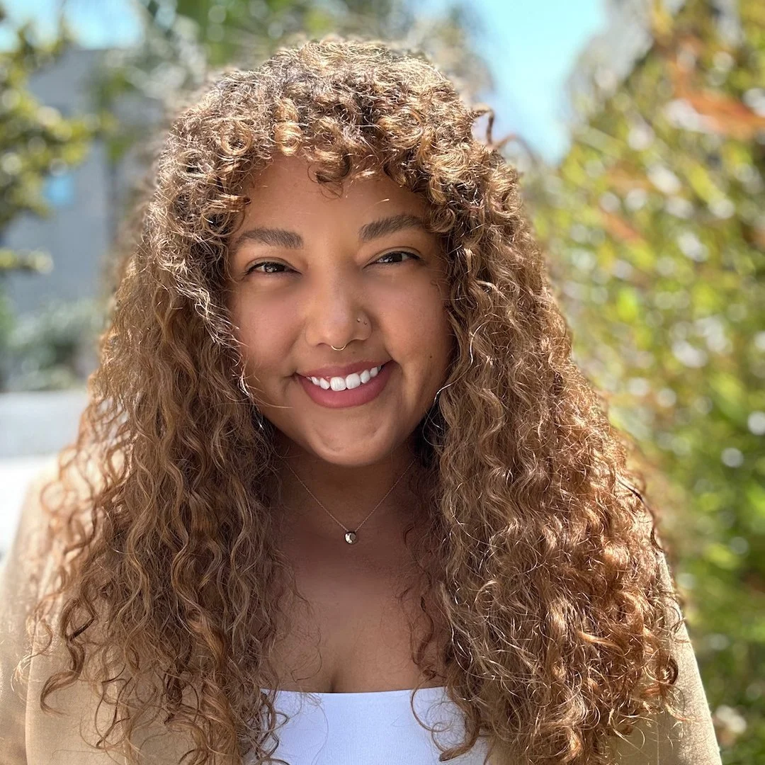 Close-up of a young woman with curly hair, smiling outdoors with trees and blue sky in the background.