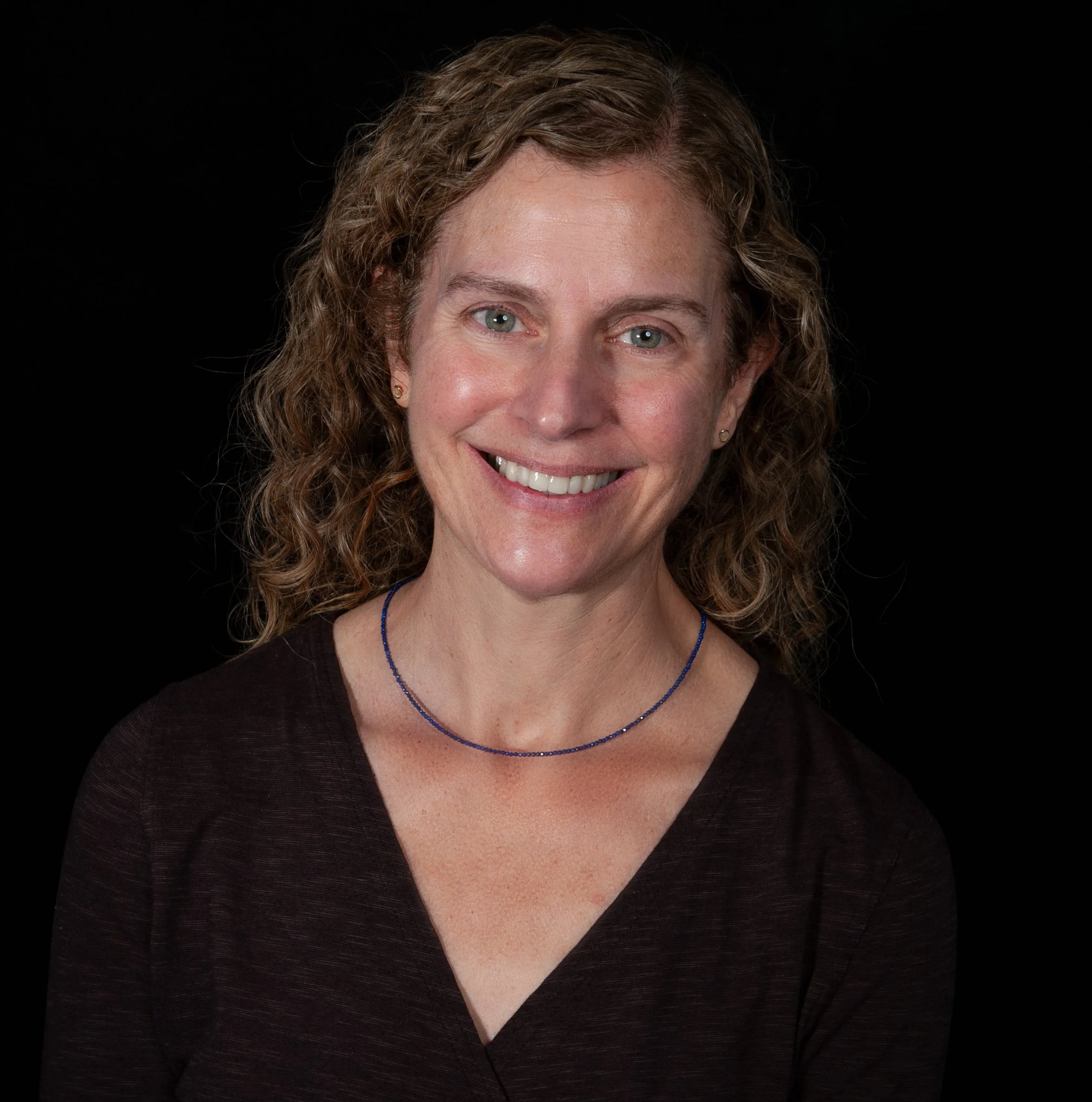 A woman with curly brown hair, blue eyes, wearing a white blouse, a pearl necklace, and earrings, smiling in front of a gray background.