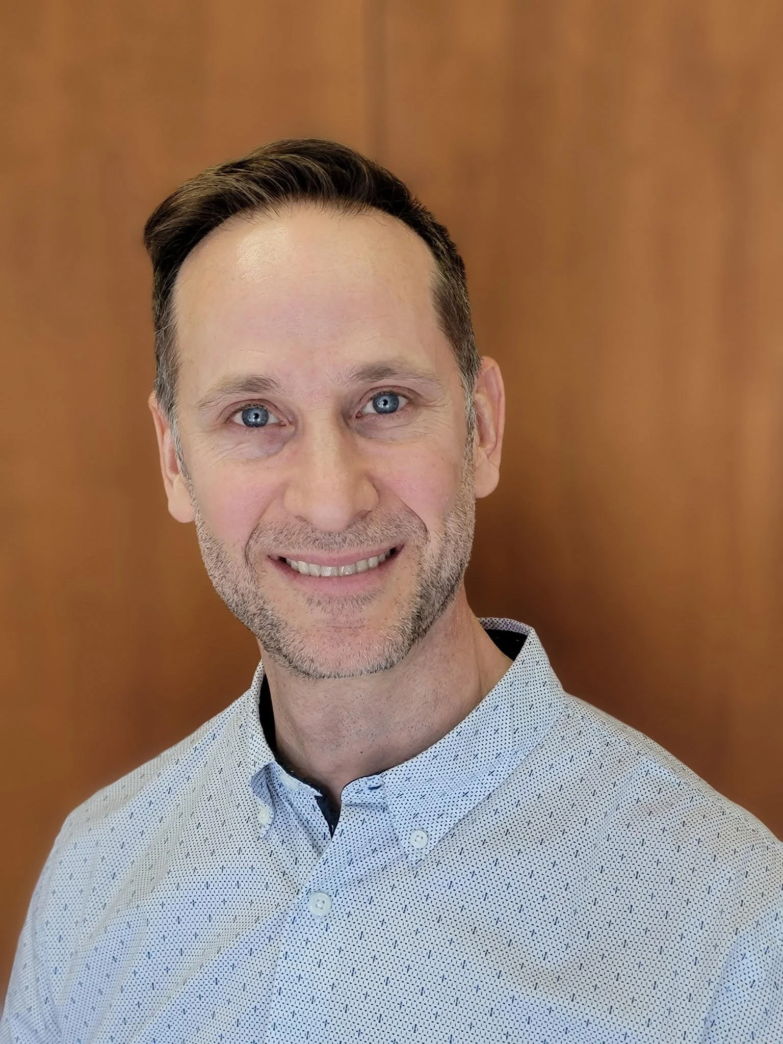 A middle-aged man with gray hair, glasses, and a goatee smiling at the camera. He is dressed in a black suit, white shirt, and a colorful tie. The background shows a warmly lit room with a lamp and a colorful painting.