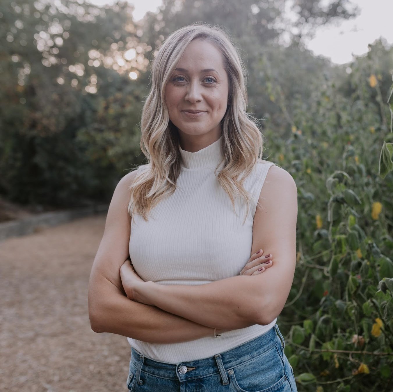 A young woman with blonde wavy hair standing outdoors with her arms crossed, wearing a sleeveless white turtleneck top and blue jeans, smiling at the camera with trees and bushes in the background.
