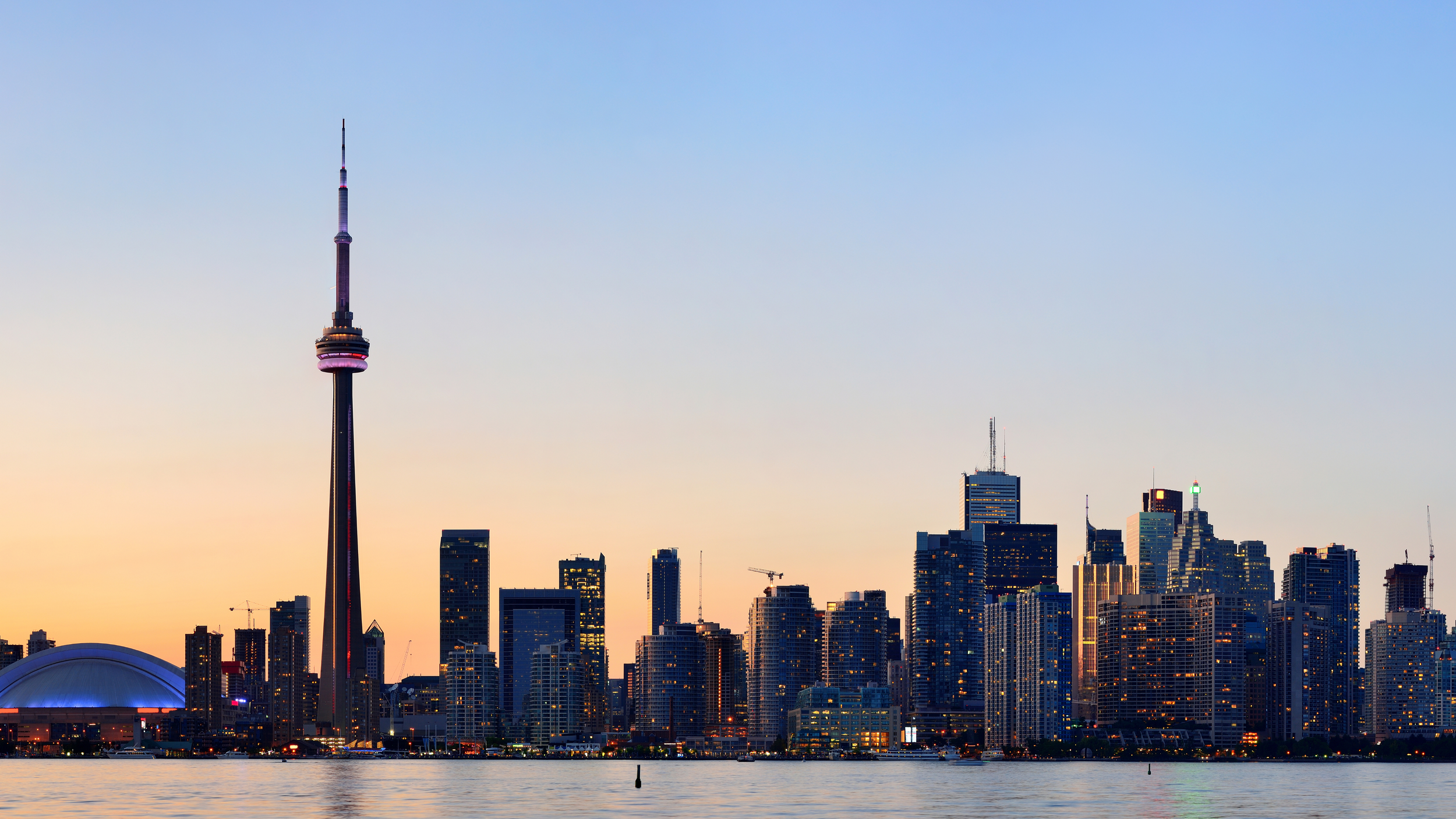Skyline of Toronto at sunset featuring the CN Tower, with tall skyscrapers and a body of water in the foreground.