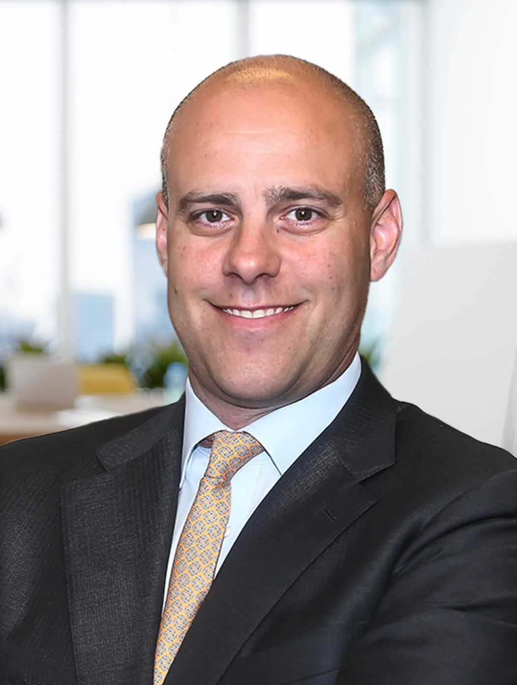 A professional man in a dark suit with a patterned tie, smiling with crossed arms in an office setting.