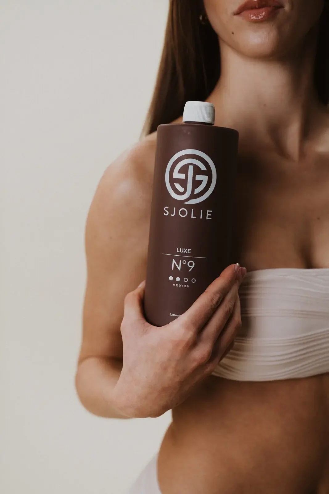 A woman holding a bottle of SJOLIE Luxe No. 9 skincare product near her shoulder.