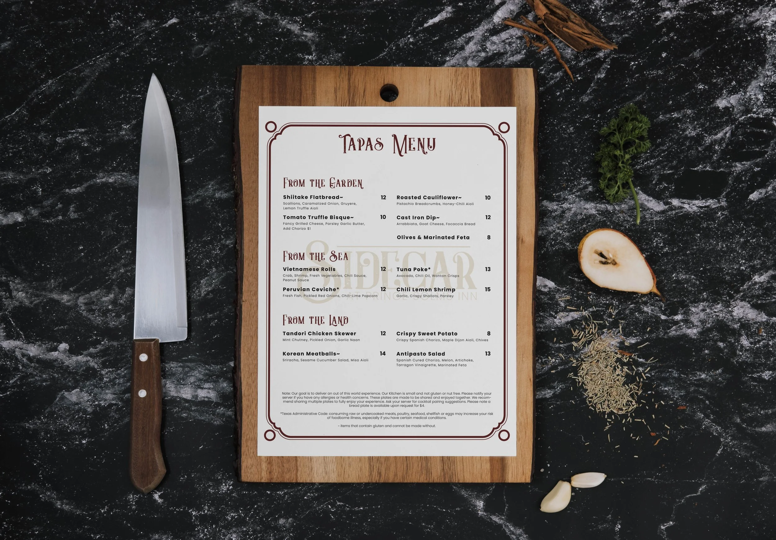A tapas menu displayed on a wooden board, featuring sections 'From the Garden,' 'From the Sea,' and 'From the Land' with various dishes. The board is placed on a dark marble surface alongside a kitchen knife, herbs, spices, half a pear, and garlic cloves.