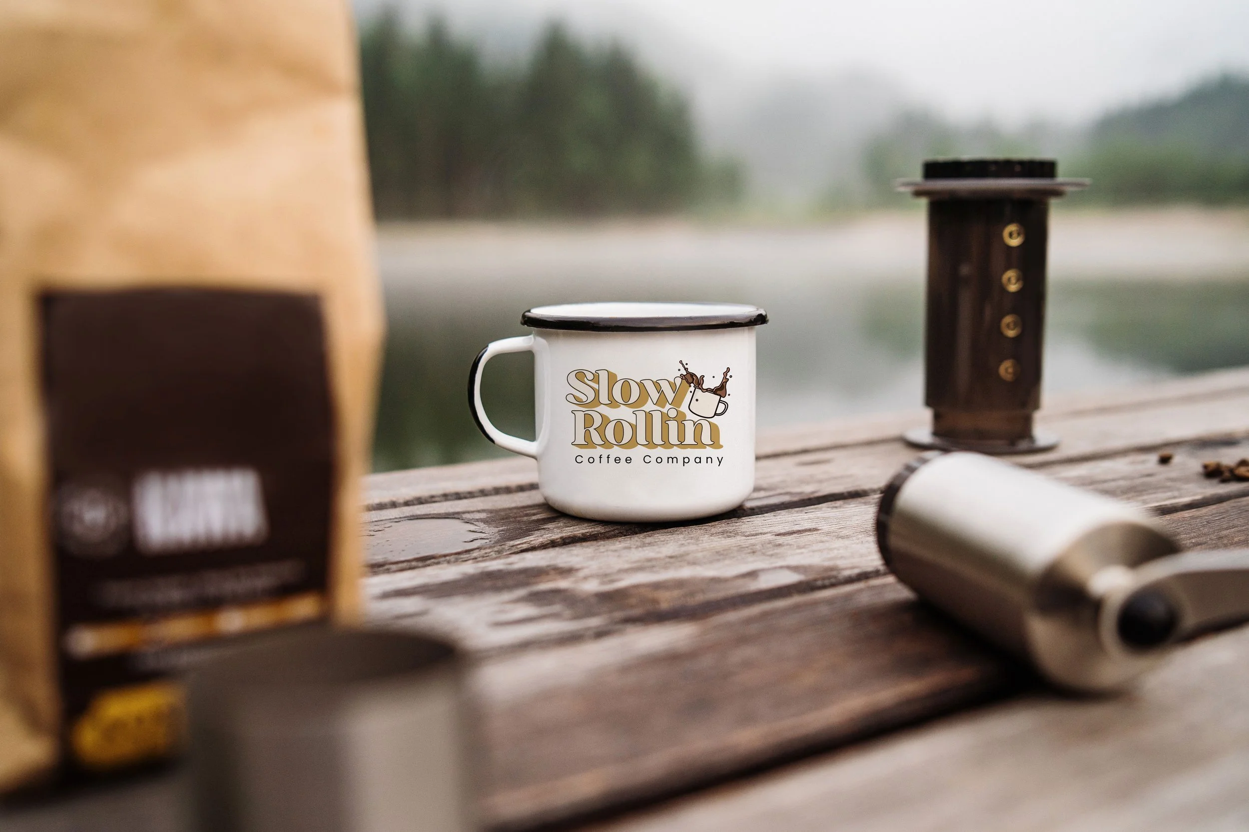 Coffee setup with a "Slow Rollin Coffee Company" mug, coffee bag, manual grinder, and AeroPress on a wooden table outdoors with a blurred natural background.