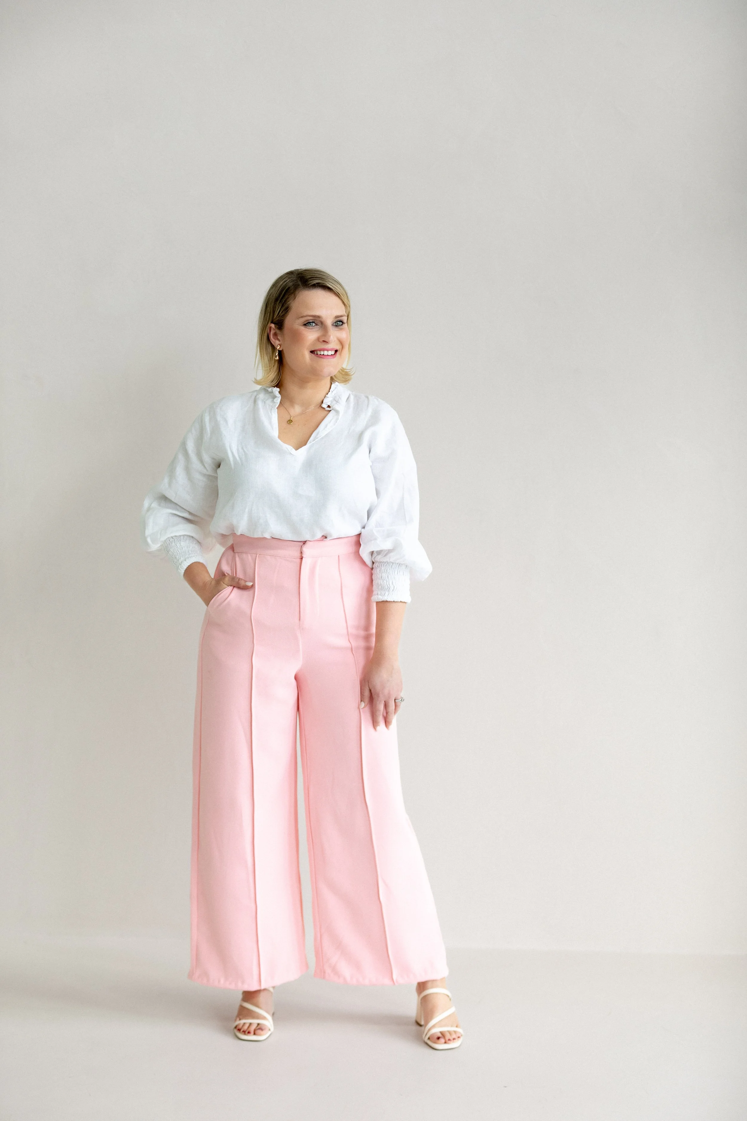 A woman with blonde hair wearing a white blouse and pink wide-leg trousers standing in front of a plain light-colored wall.