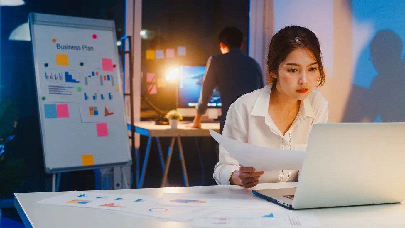 A woman working on a laptop at a desk with business reports and charts, in an office with a whiteboard and a man working on a computer in the background.