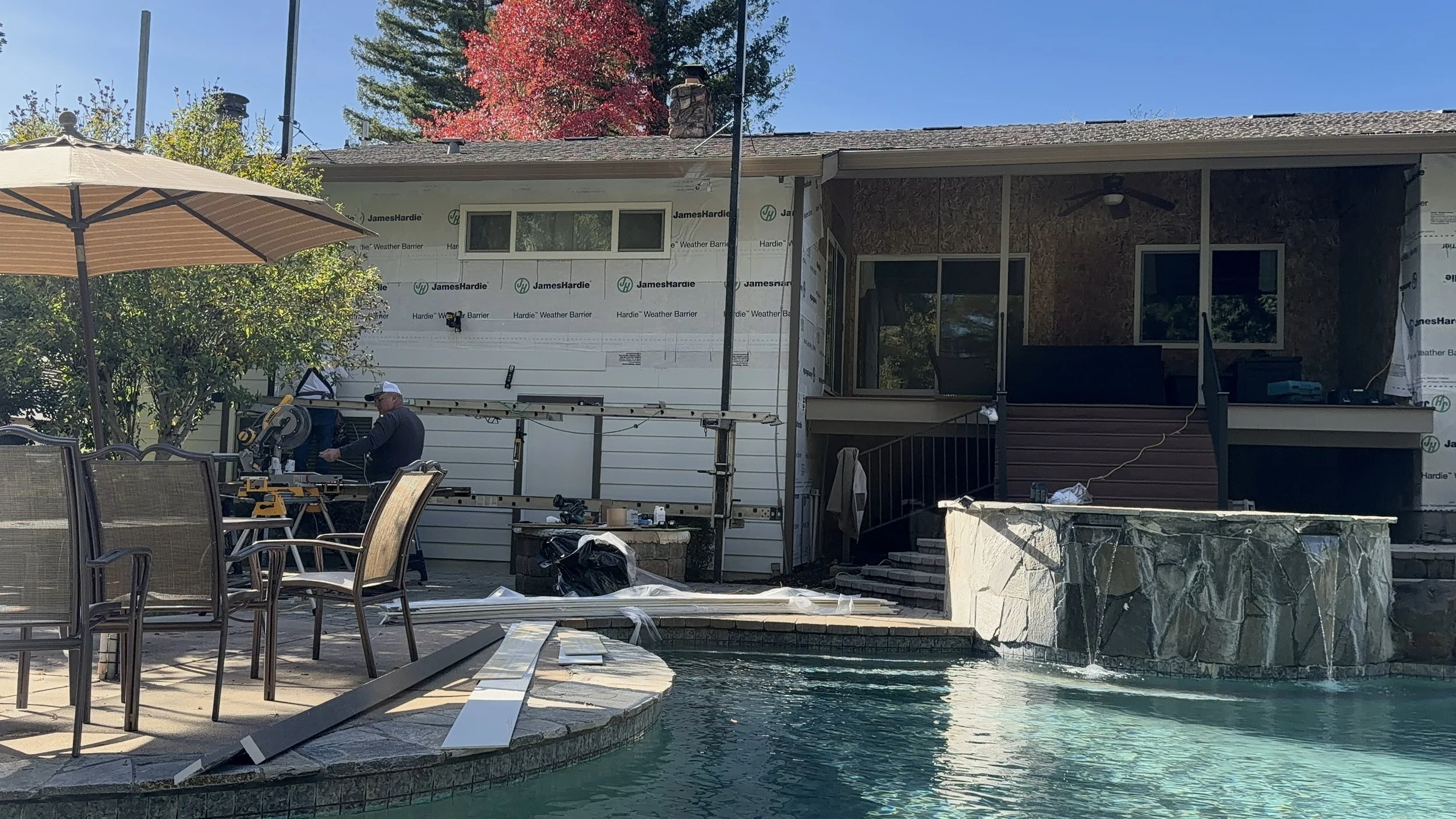 Construction workers building a house backyard patio with a swimming pool, tables, chairs, umbrella, and a stone waterfall feature.