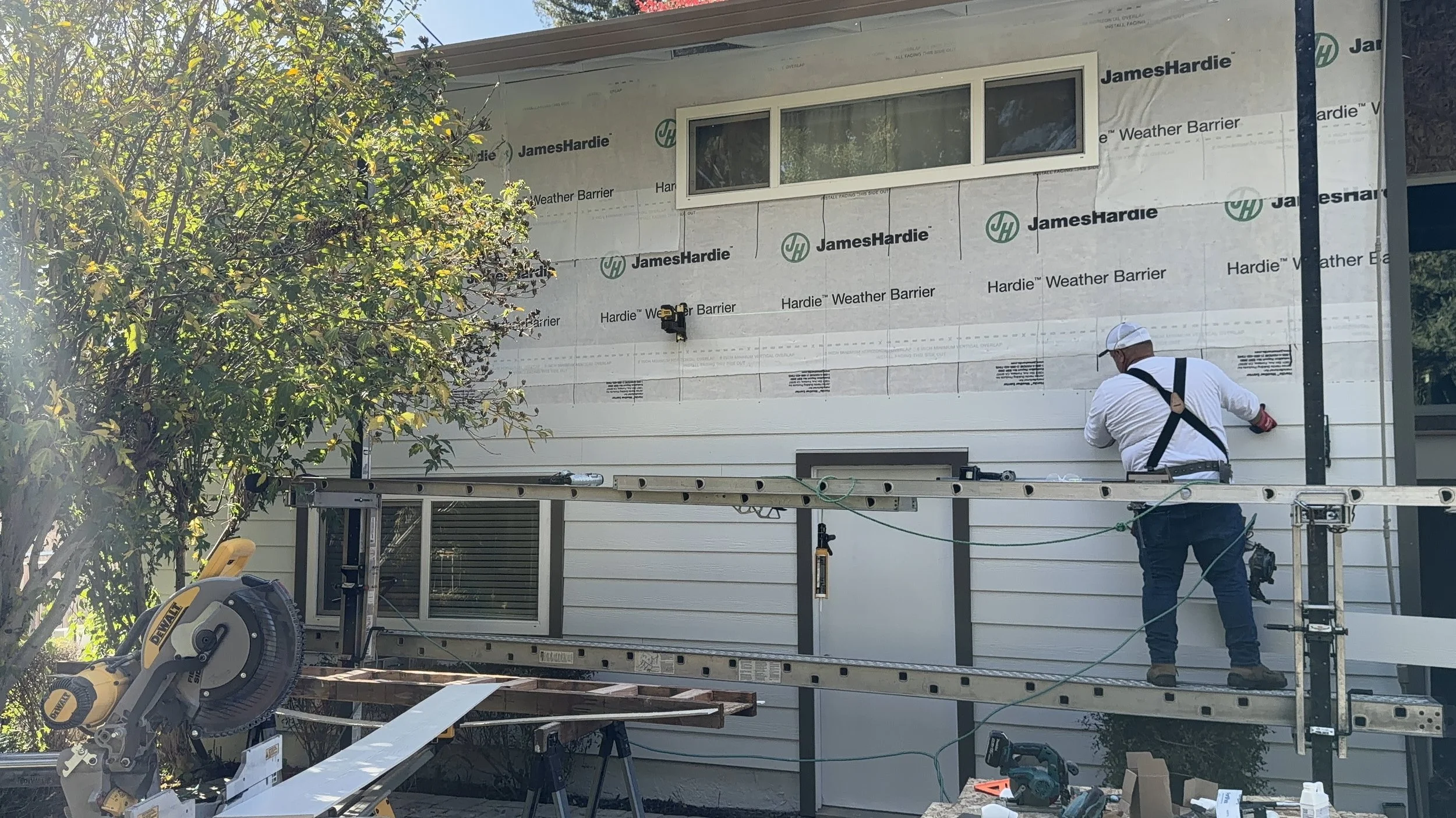 A construction worker on scaffolding installing siding on the exterior of a house, with a tree and construction tools in the foreground.