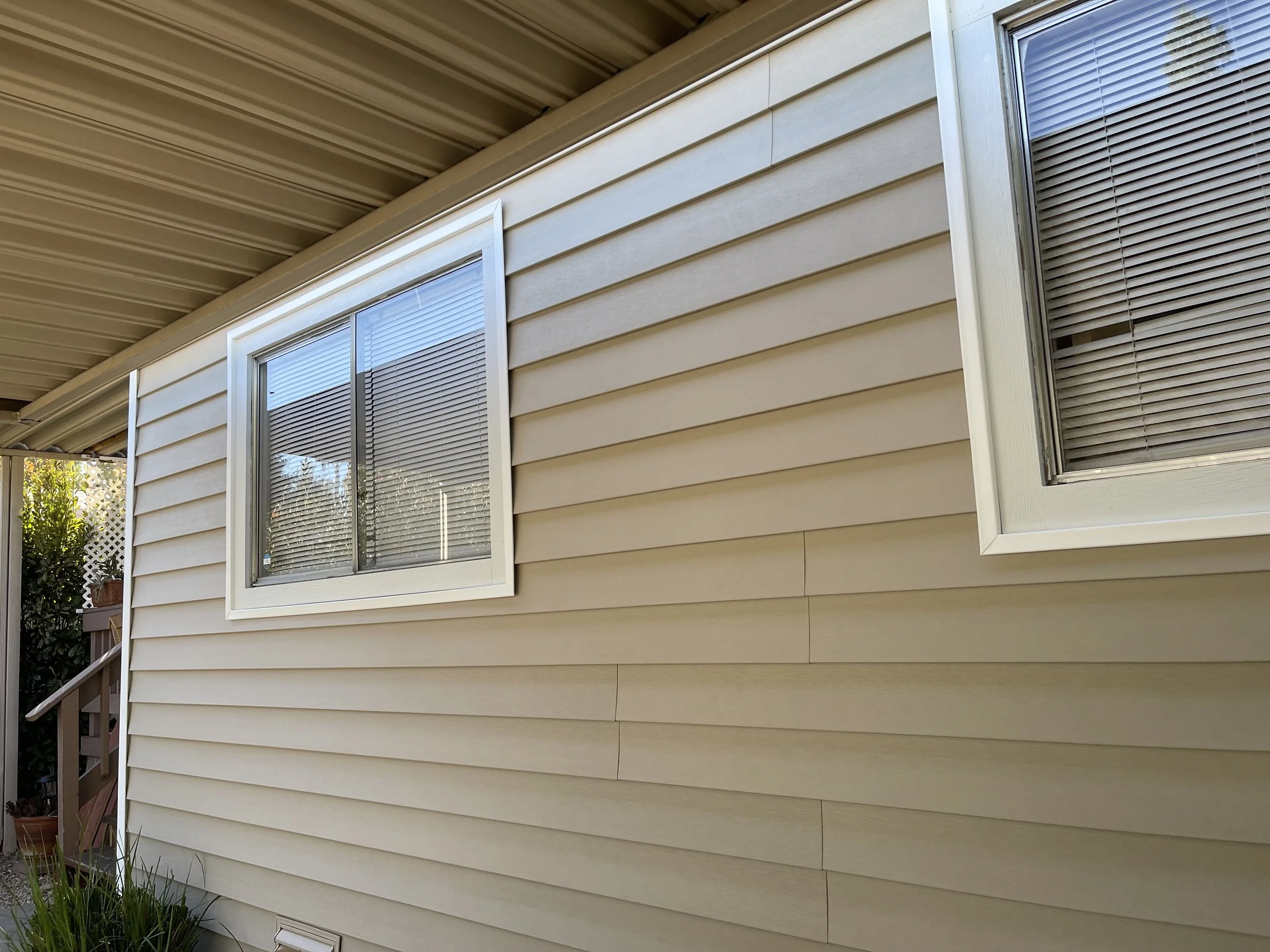 Exterior view of a beige house with vinyl siding, showing two double-pane windows with blinds, located under a sloped metal roof.