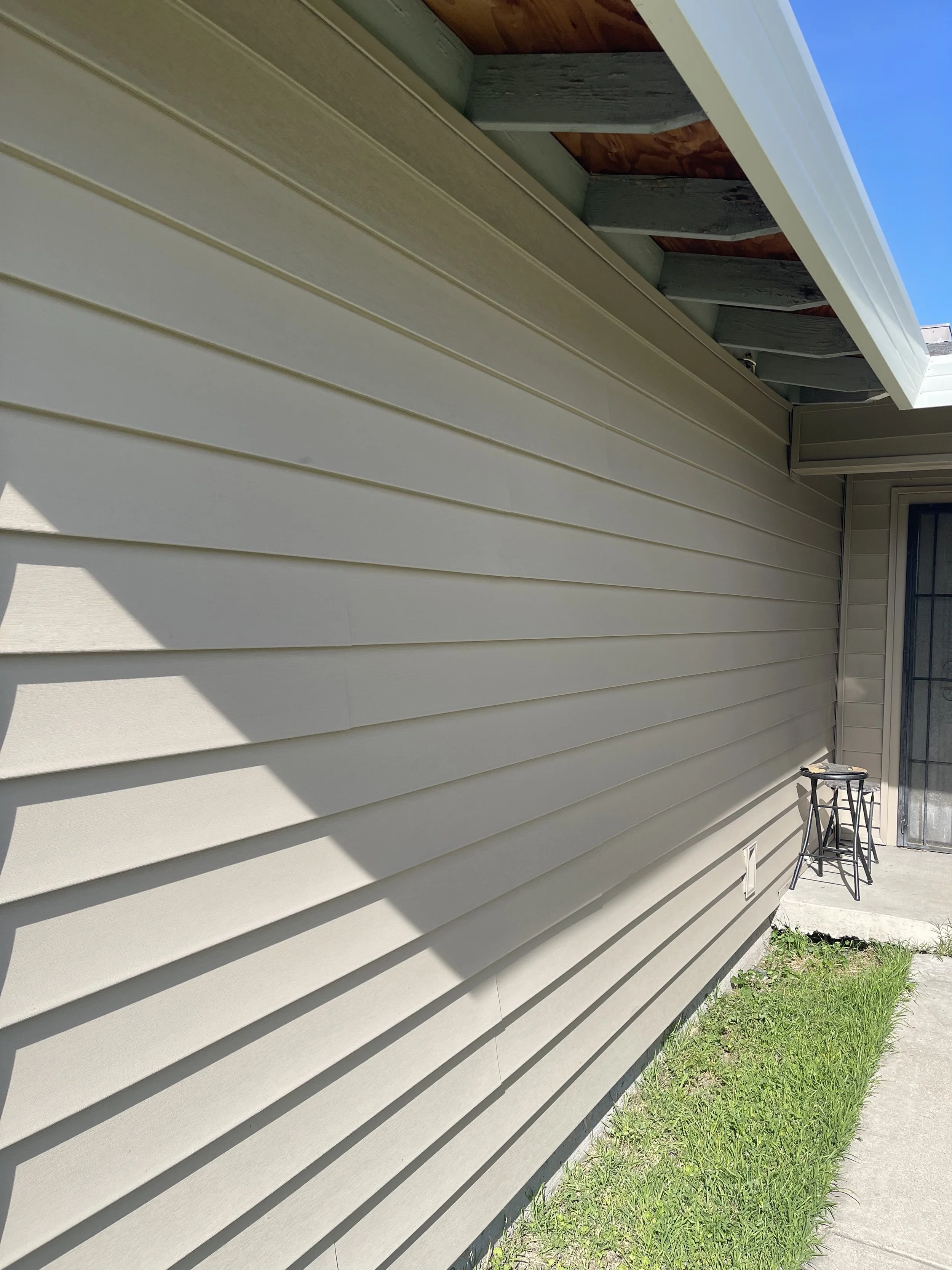 Exterior beige siding wall of a house with a shadow cast on it, next to a small concrete patio with two stool chairs and a glass sliding door, grass in the foreground, partly cloudy sky above.