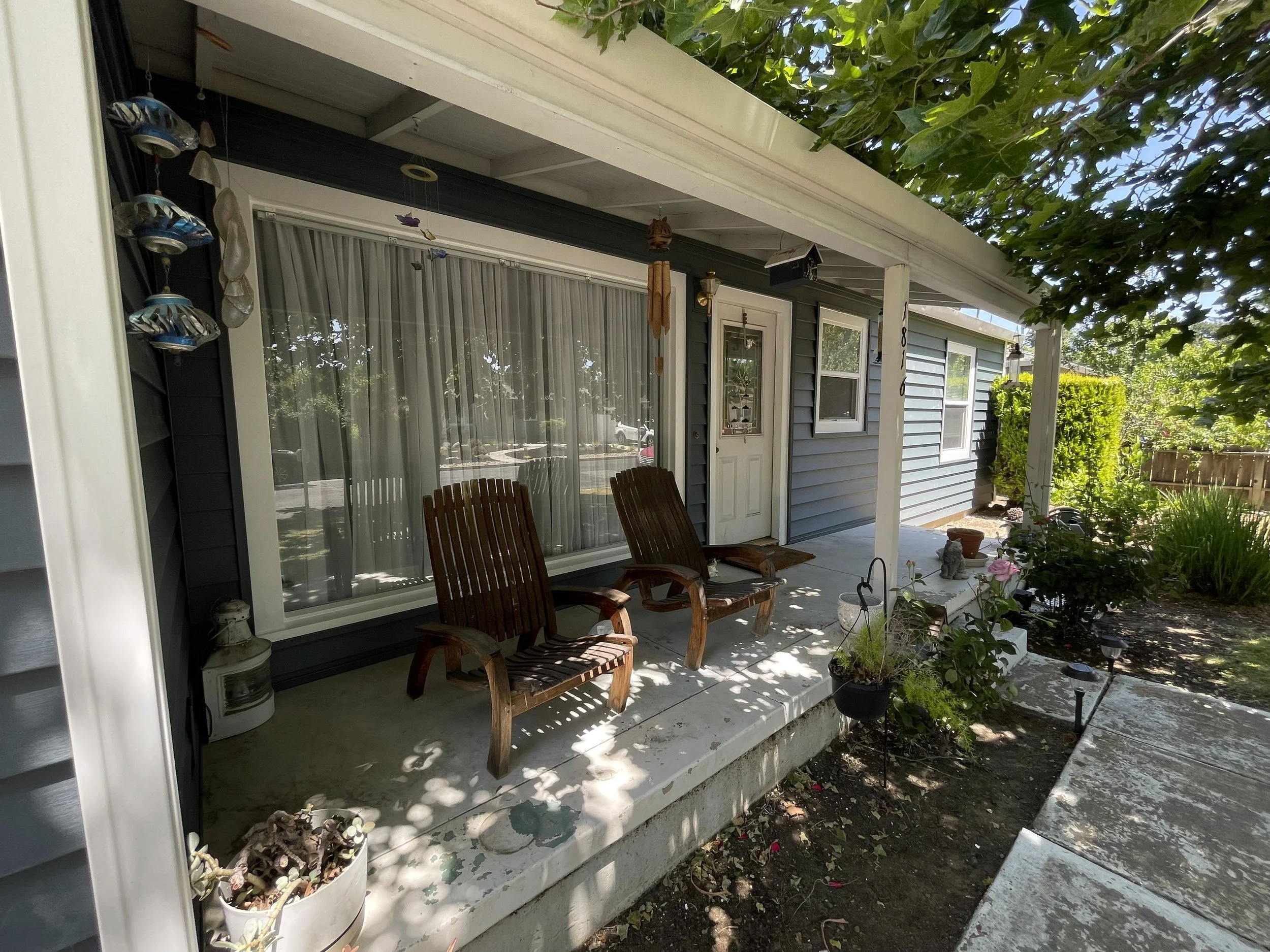 Front porch of a house with two wooden chairs, potted plants, hanging decorations, and a small garden area.