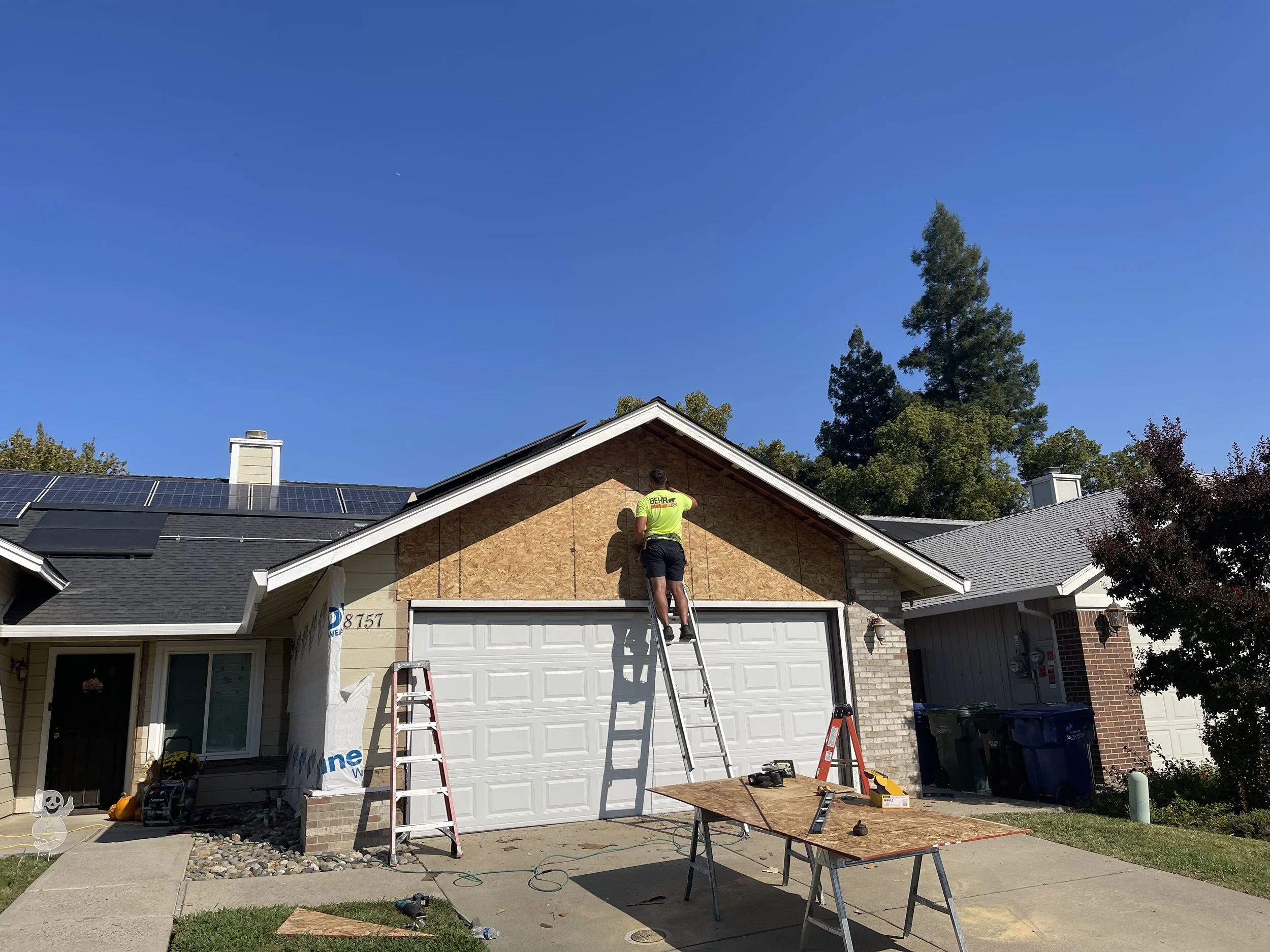 A man wearing a neon green shirt and black shorts is working on the front of a house during the day. He is standing on a ladder, installing or repairing the upper part of the house's facade, which is covered with plywood. There are tools and construc