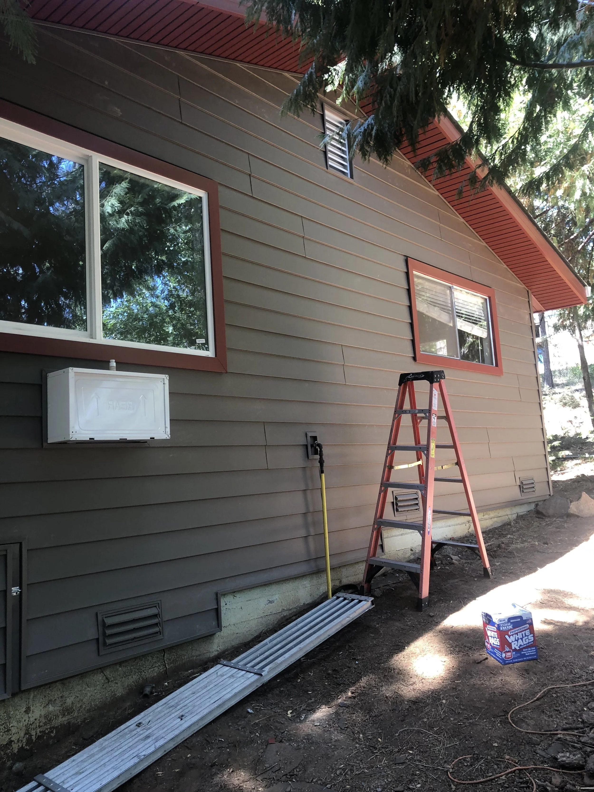 Side view of a house with brown siding, two windows, a ladder, a yellow broom, a box of white rags, and some construction debris on the ground.