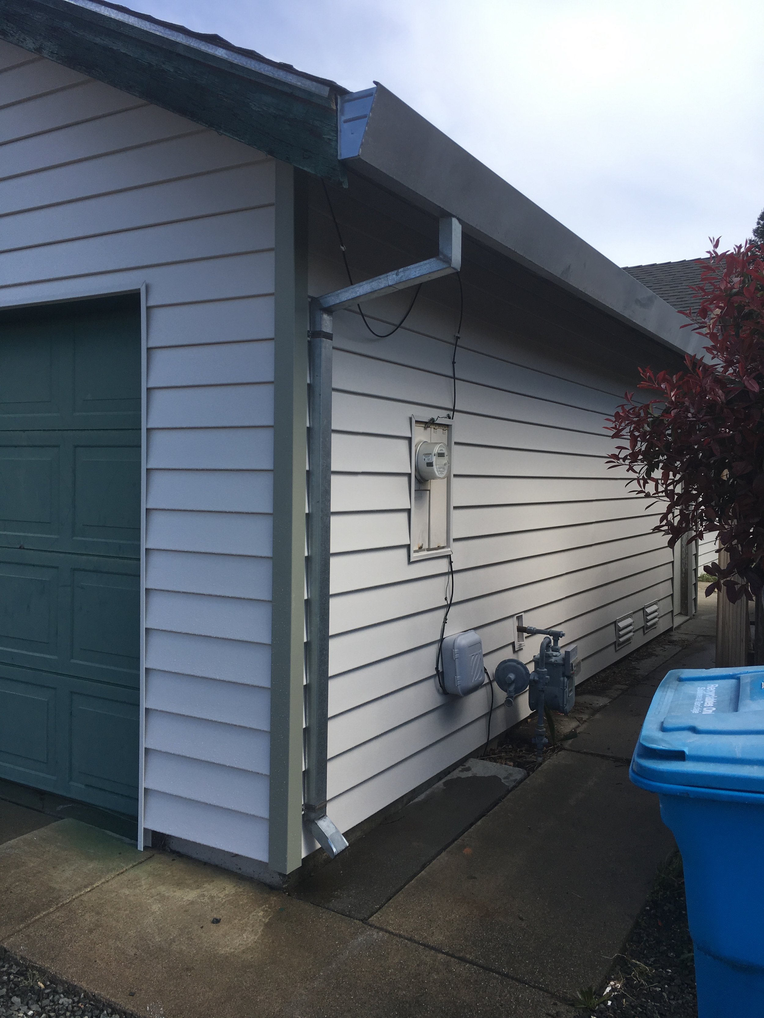 Side of a house with beige vinyl siding, a green garage door, an electrical meter, and outdoor pipes, with a blue trash bin in the foreground.