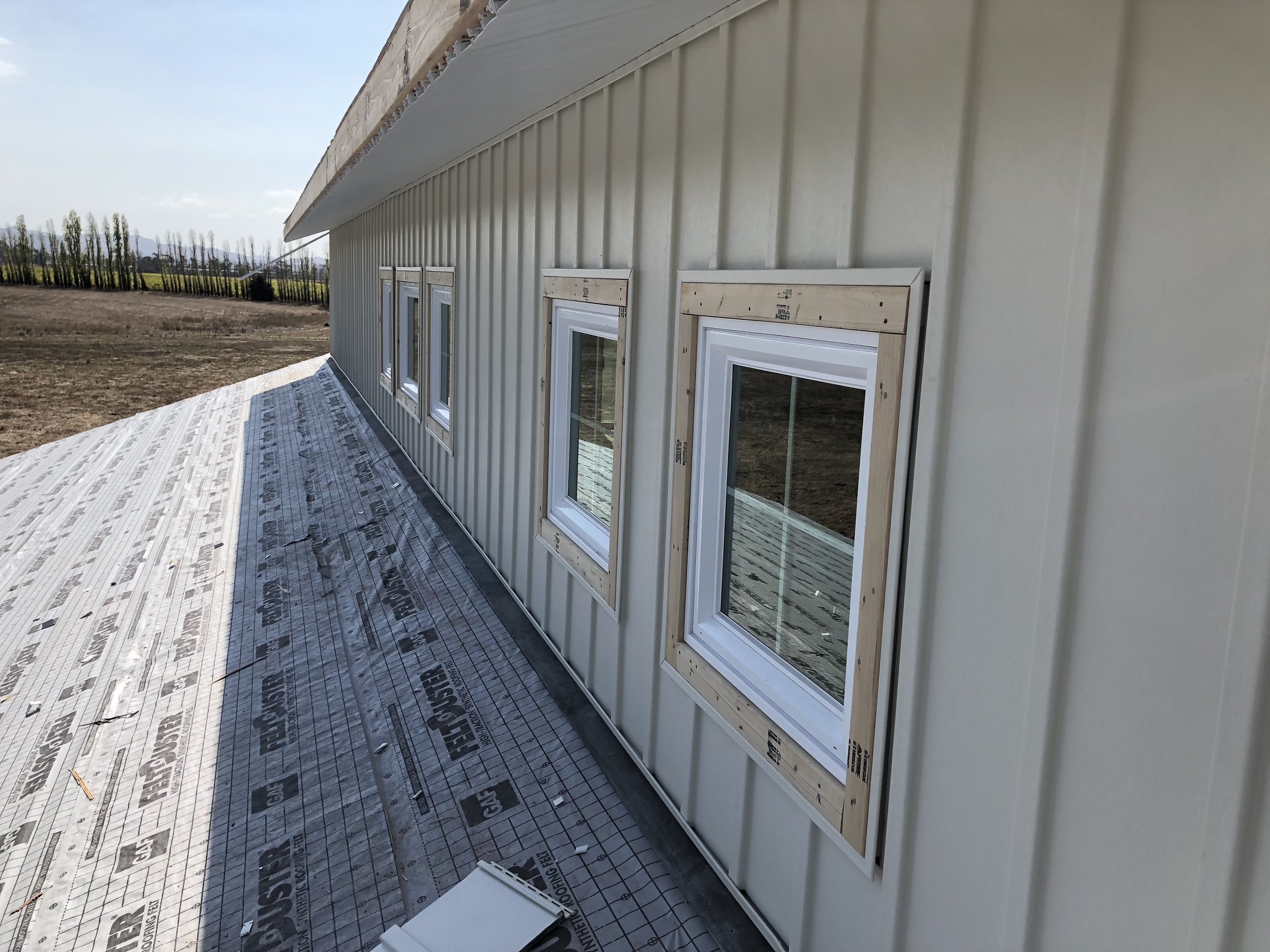 Building exterior with four installed white-framed windows on a metal siding wall, with a sloped metal roof, on a construction site surrounded by open land.