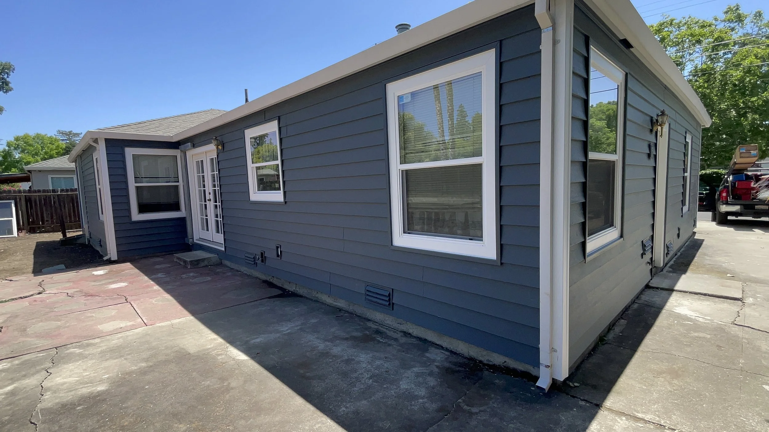 Blue house with white trim, multiple windows, and a patio with cracks, under a clear blue sky.