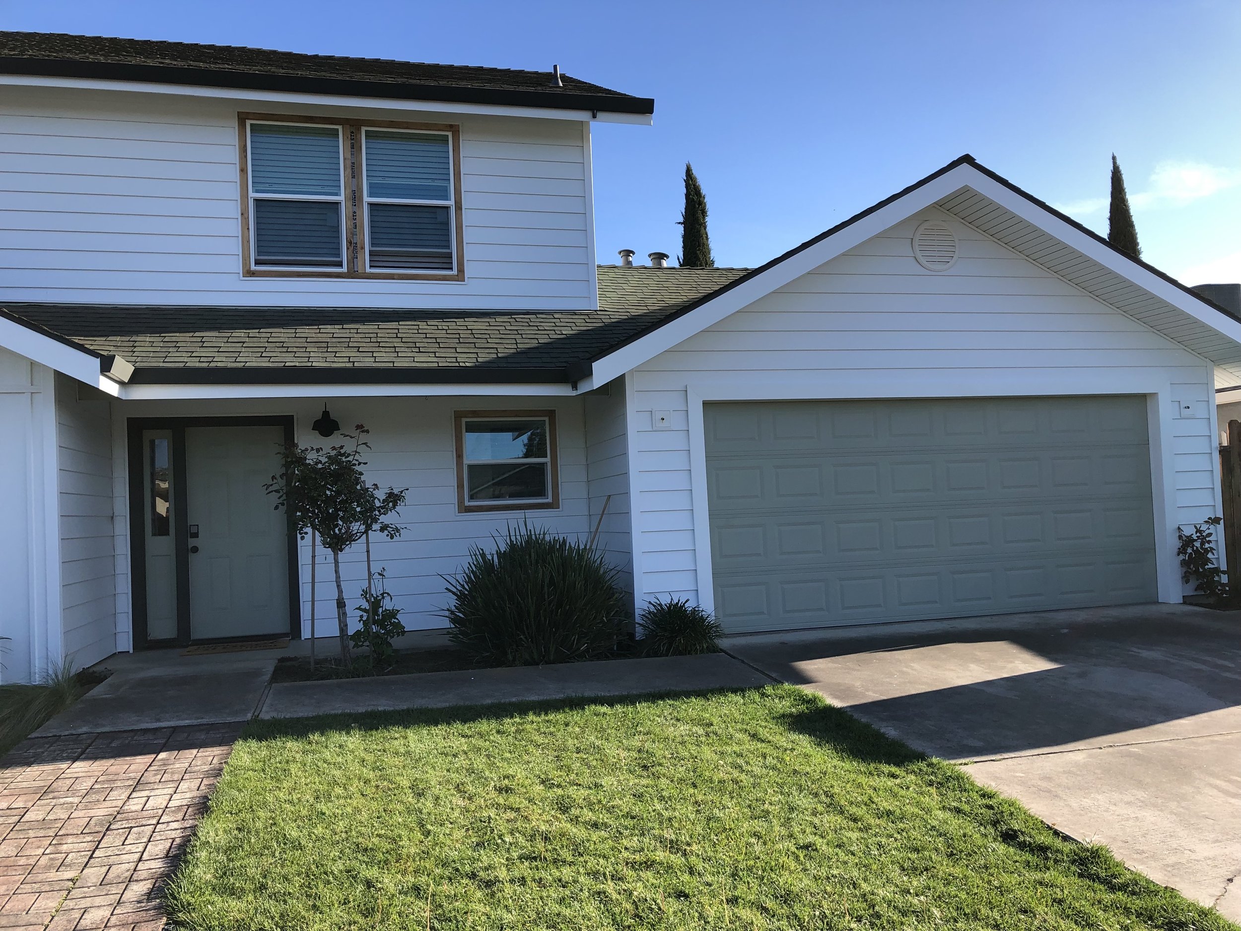 Front view of a two-story house with a white exterior, gray front door, attached garage, small front yard with grass and plants, and a clear blue sky.