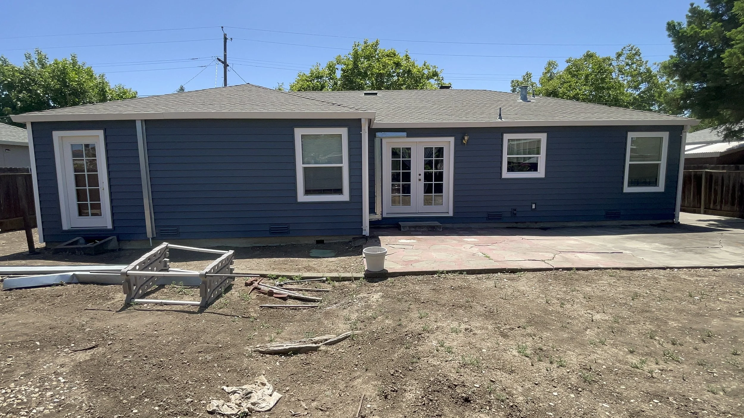 Backyard view of a house with blue siding, two sets of French doors, four windows, and a new concrete patio. The yard is mostly bare dirt with some construction materials and tools scattered around.