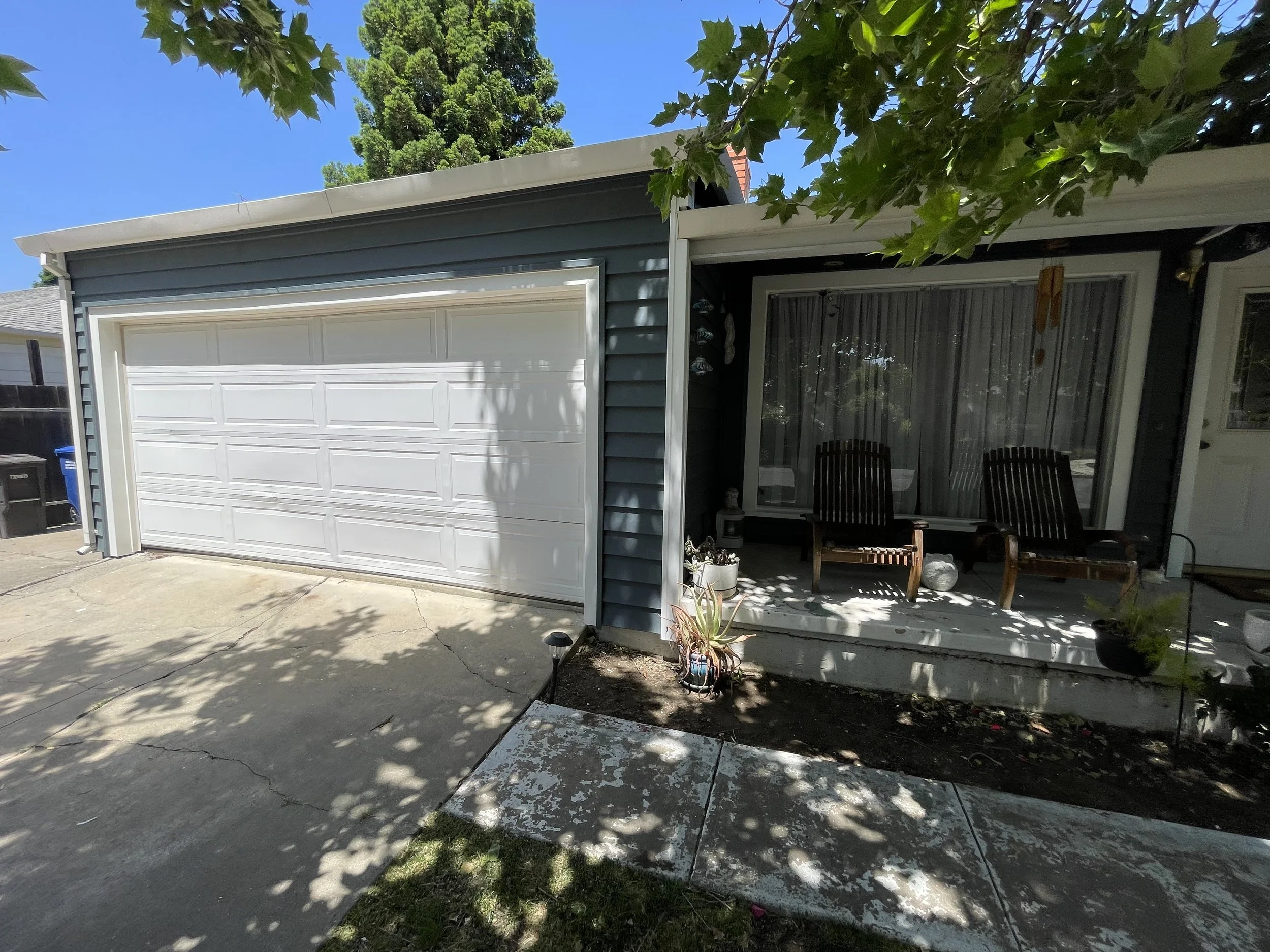 Front view of a house with a gray siding exterior, white garage door, and a small porch with two dark wood chairs, potted plants, and hanging decorations, partially shaded by tree branches.