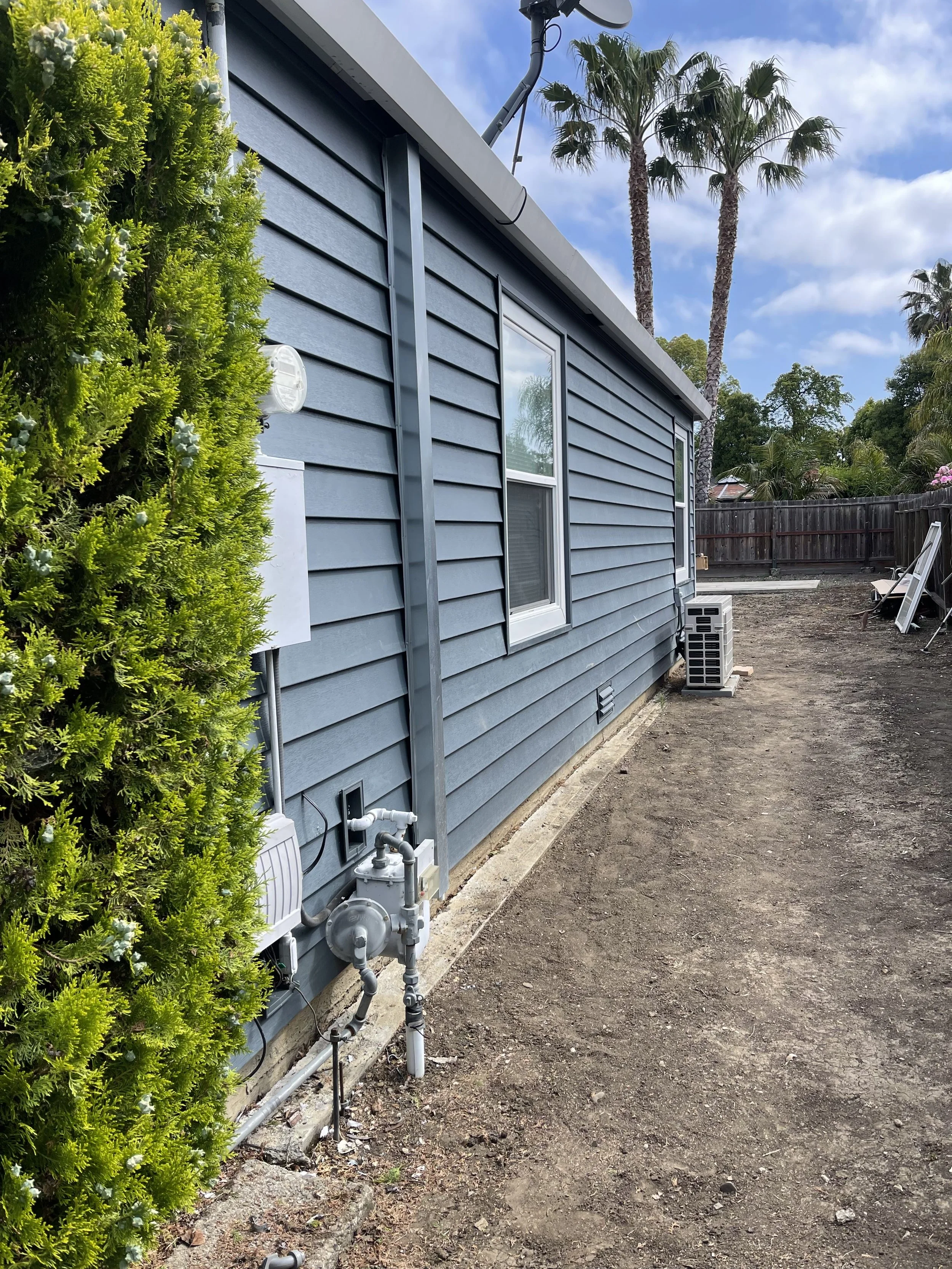 The side of a house painted in blue with two white-framed windows. There is a green shrub on the left, pipes and electrical components on the wall, and an AC unit on the ground. Palm trees and a partly cloudy sky are in the background.